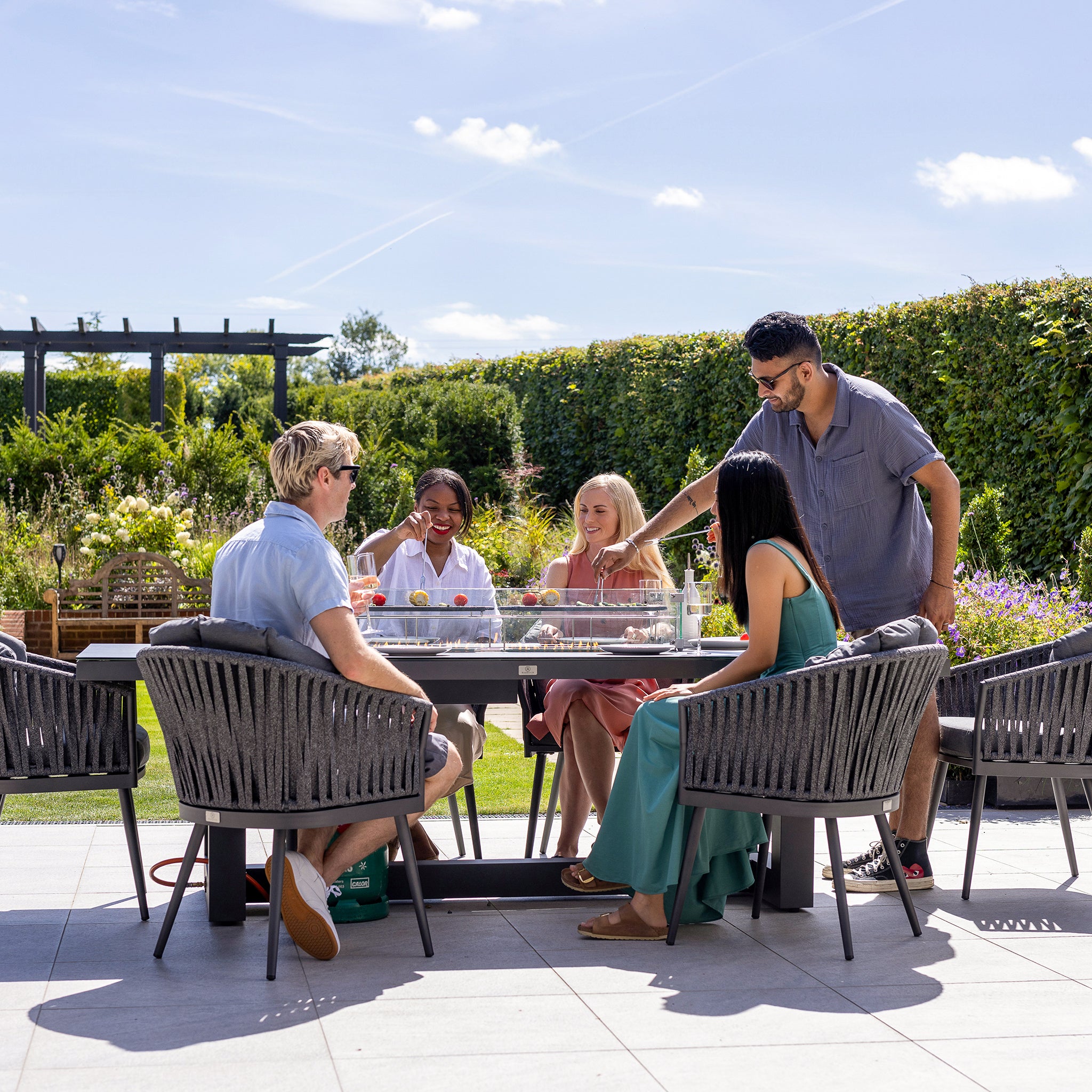 Five people sitting and standing around a Monterrey 8 Seat Rope Rectangular Ceramic Firepit Dining Set in Grey, enjoying a sunny day in a lush garden setting. The high-quality aluminum table complements the relaxed vibe of the Harbour Lifestyle Monterrey collection.