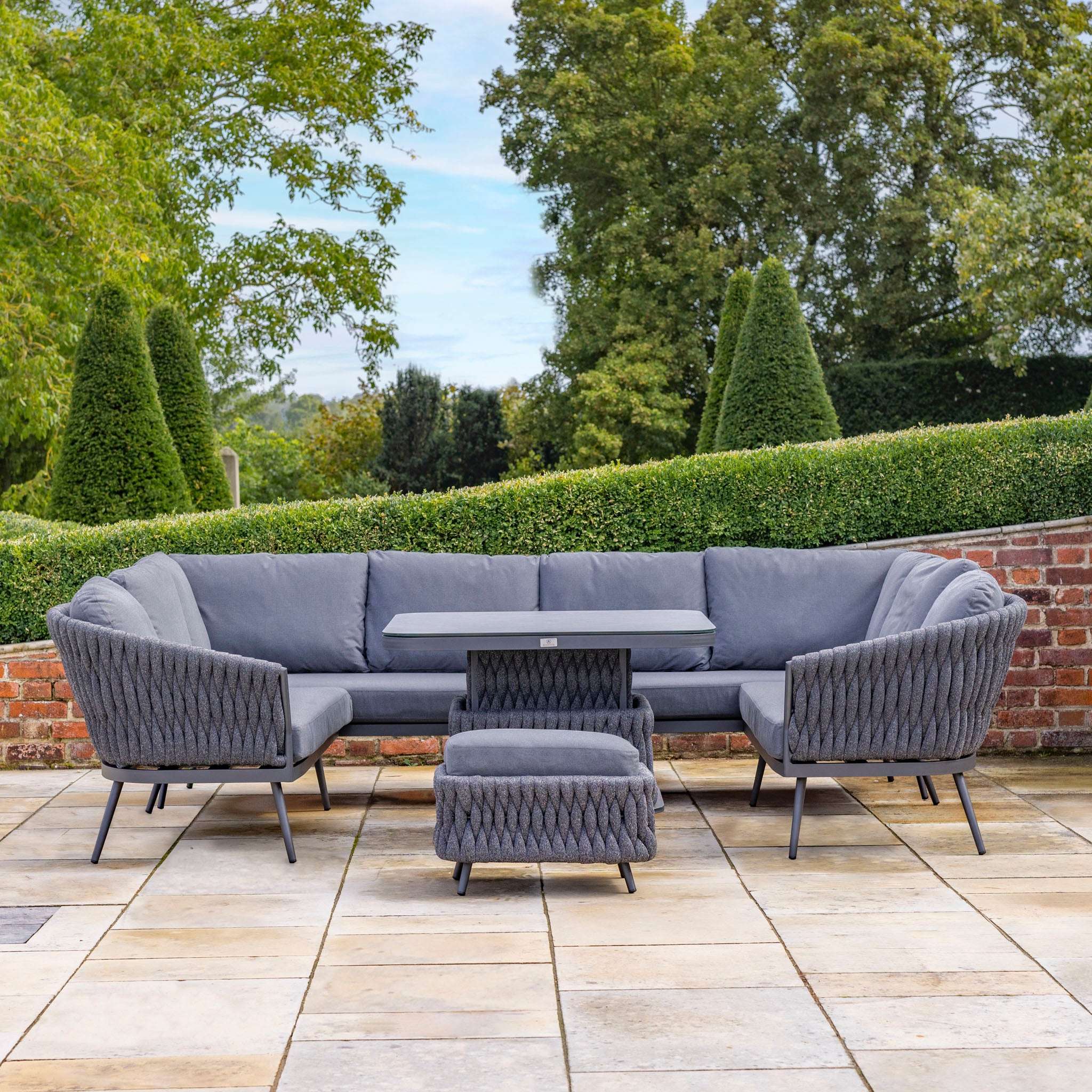 Outdoor patio set with gray, UV-resistant fabric cushioned seating, a rectangular table, and an ottoman on a stone patio amid lush greenery. Experience the Palma U-Shape Rope Corner Dining Set with Rising Table in Grey in this cozy oasis.