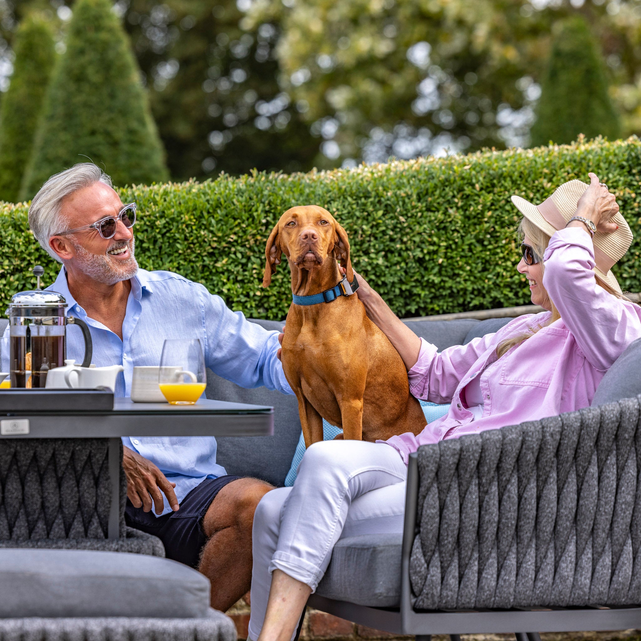 A man and a woman relax outdoors with a brown dog sitting on the table between them, surrounded by greenery, enjoying their Harbour Lifestyle Palma U-Shape Rope Corner Dining Set with Rising Table in Grey.