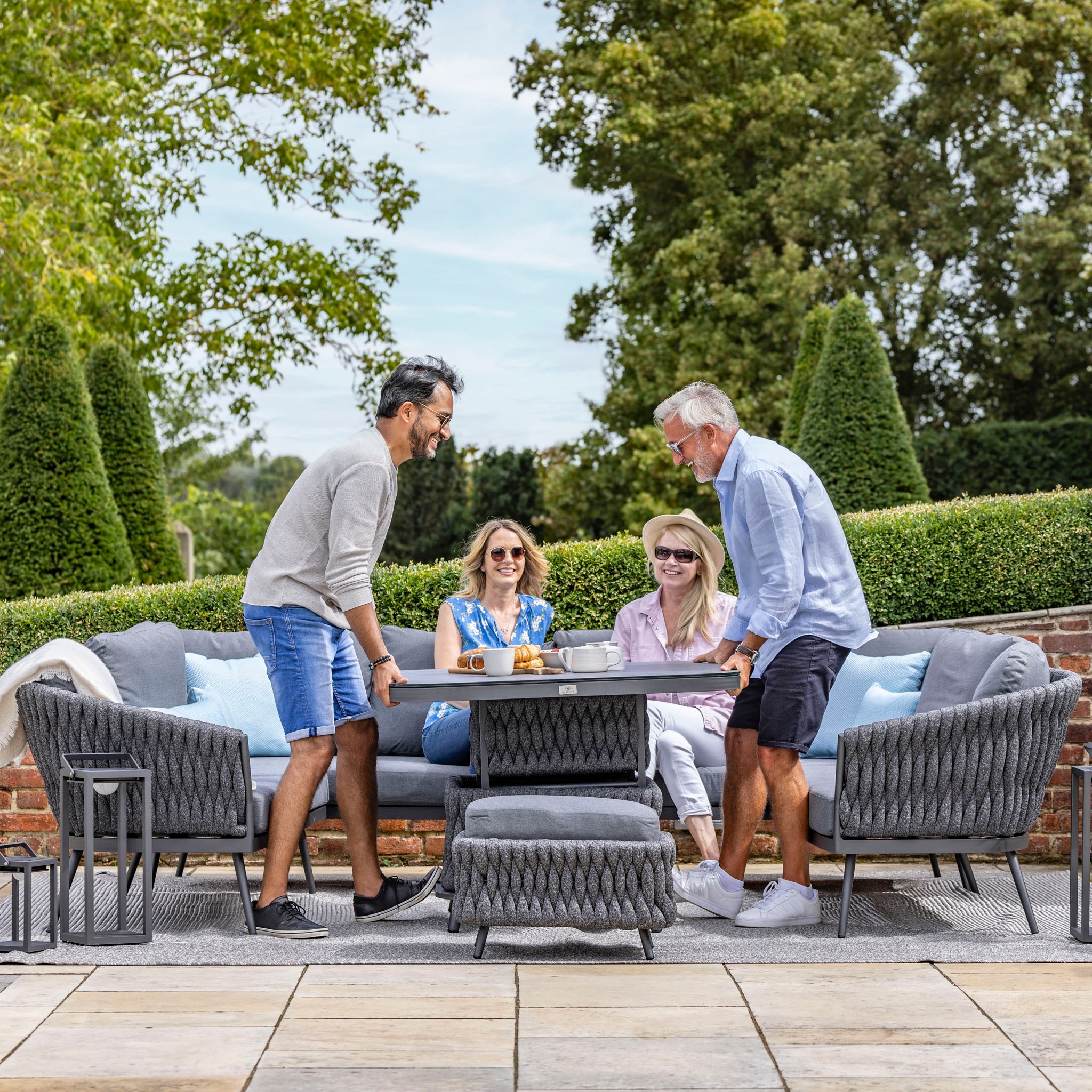 Four people enjoy a meal at an outdoor patio table, comfortably seated on the Palma U-Shape Rope Corner Dining Set with Rising Table in Grey by Harbour Lifestyle, surrounded by greenery and trees on a sunny day.