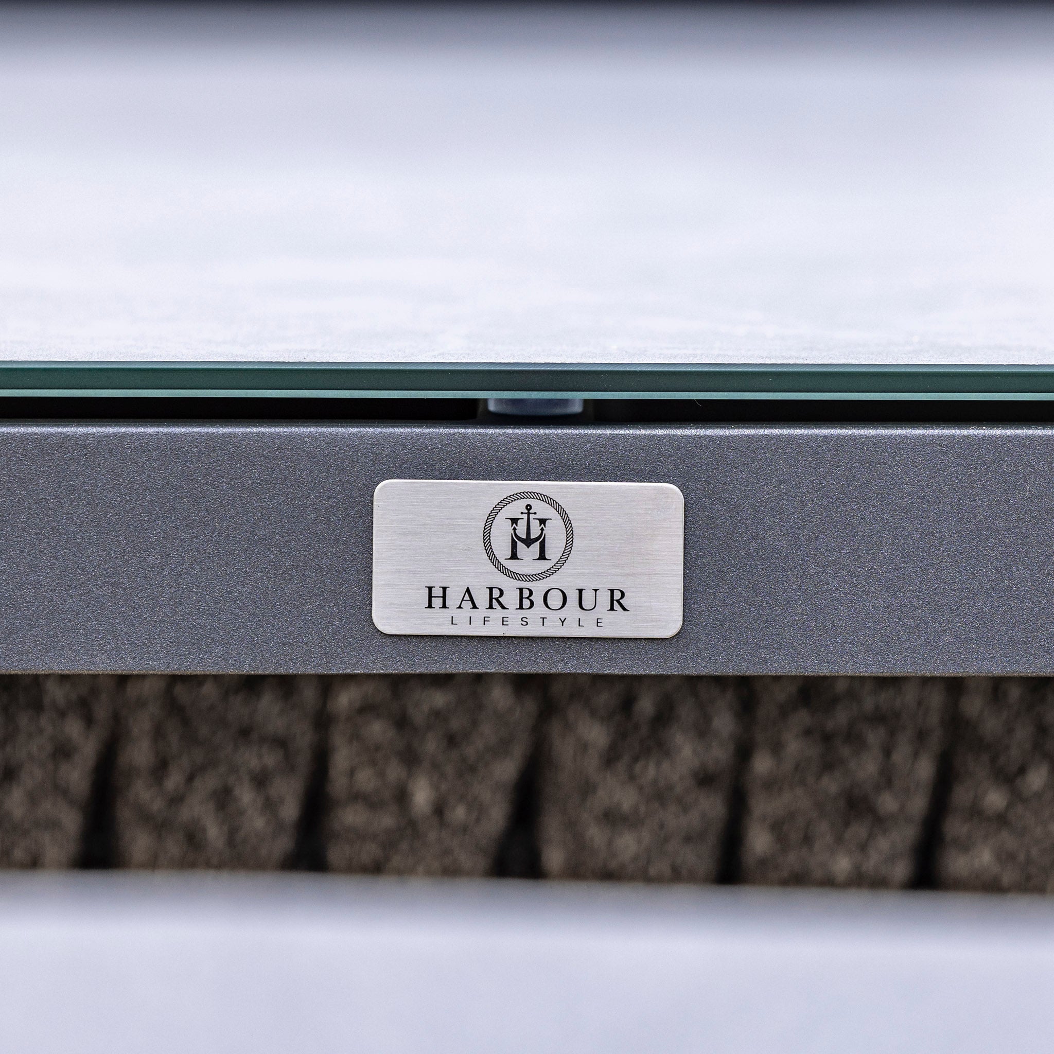 Close-up of the Harbour Lifestyle logo on a metal surface. The logo, featuring an anchor within a circle above the brand name, complements their Palma Rope Rectangular Corner Dining Set with Rising Firepit Table in Grey (Right Hand), with its aluminium frames and water-repelling thick rope weave.