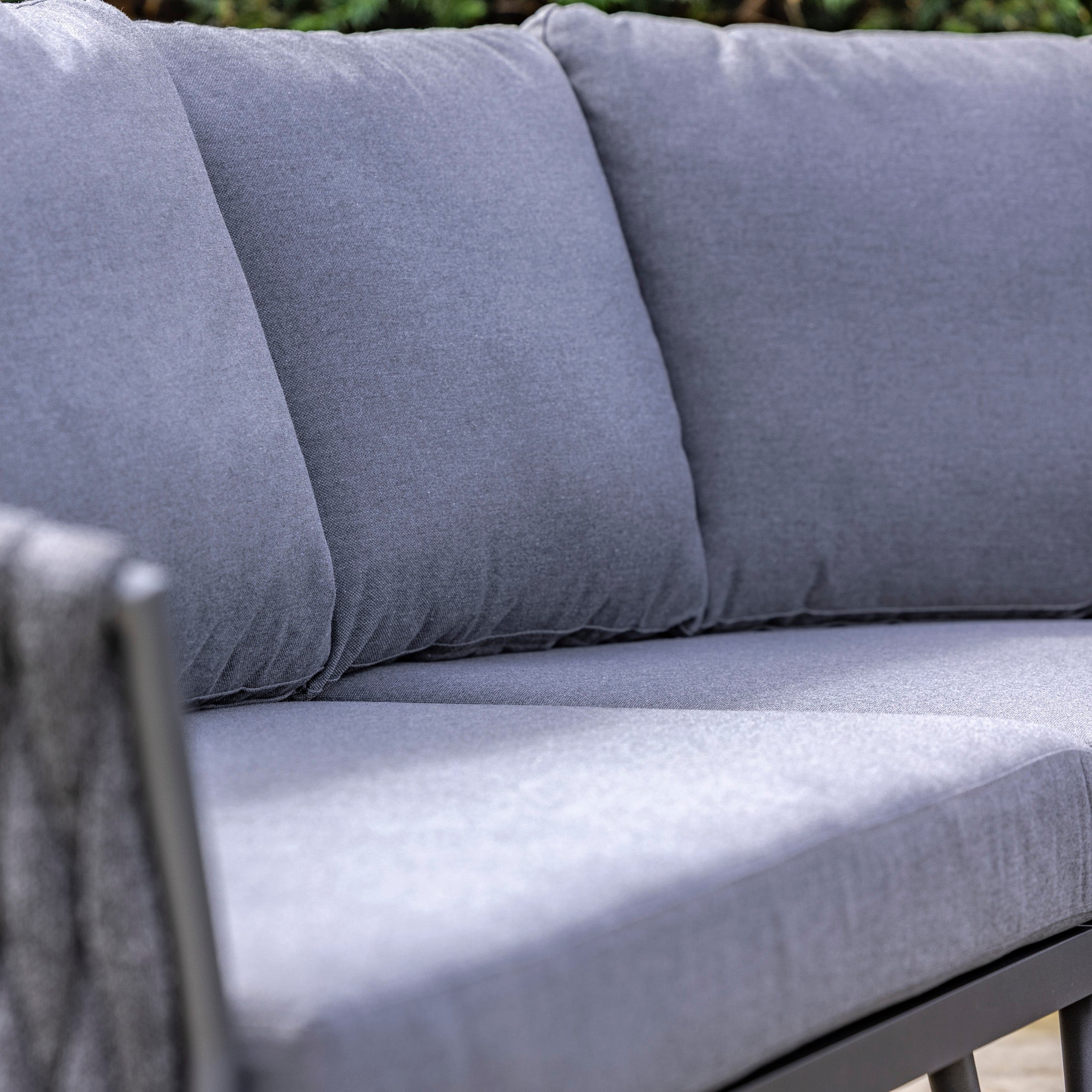Close-up of the Palma Rope Rectangular Corner Dining Set with Rising Firepit Table in Grey (Right Hand), featuring a gray cushioned outdoor sofa with a water-repelling thick rope weave, set against a leafy background.