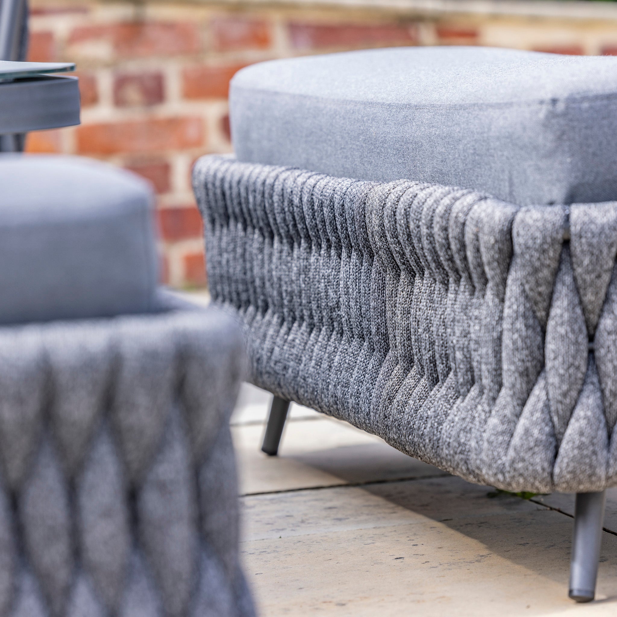 Close-up of a gray upholstered ottoman with textured fabric and black legs, placed on a tiled patio beside the Palma Rope Rectangular Corner Dining Set with Rising Firepit Table in Grey (Right Hand), featuring its water-repelling thick rope weave and sturdy aluminium frames, set against a brick wall background.