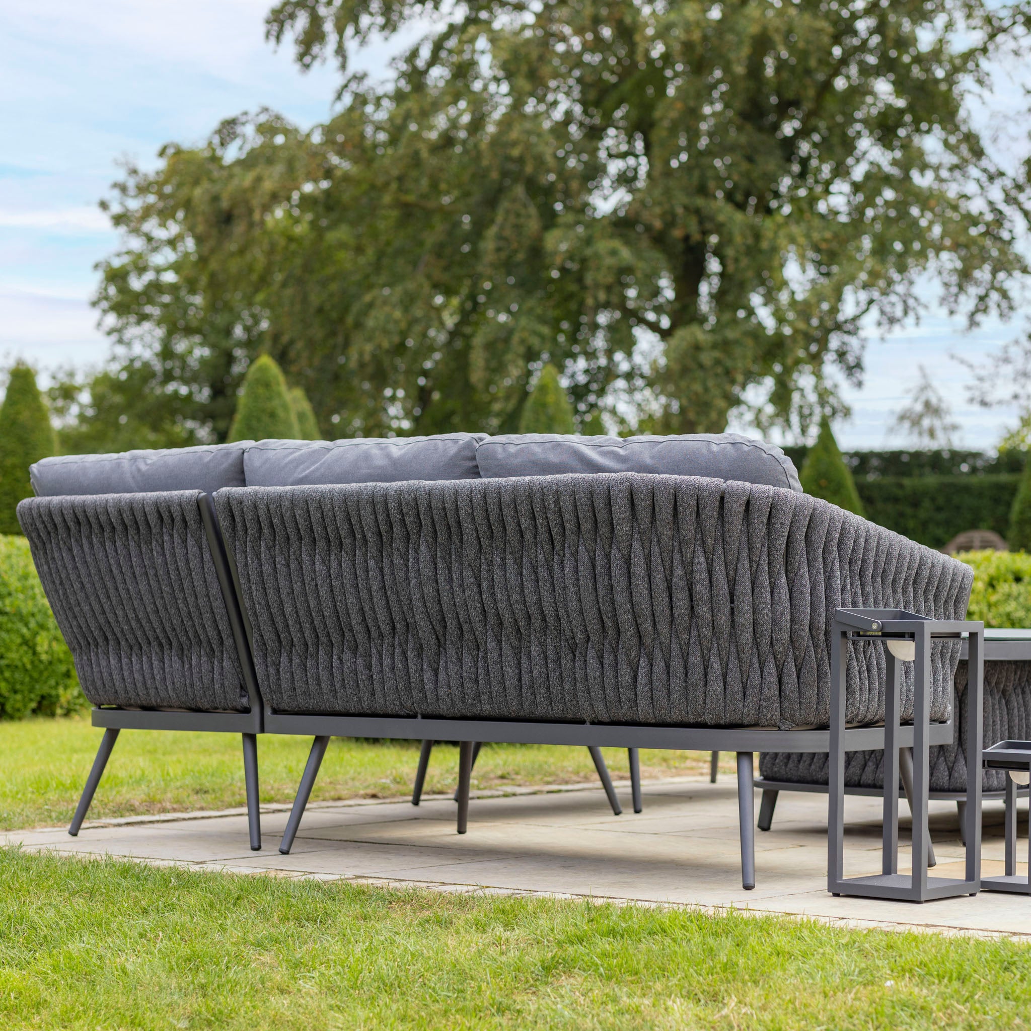 A cozy outdoor seating area featuring a gray cushioned sofa with sturdy aluminium frames and a small black table on the patio, surrounded by lush greenery. The setup is reminiscent of the elegant Palma Rope Rectangular Corner Dining Set with Rising Firepit Table in Grey (Right Hand), known for its water-repelling thick rope weave.
