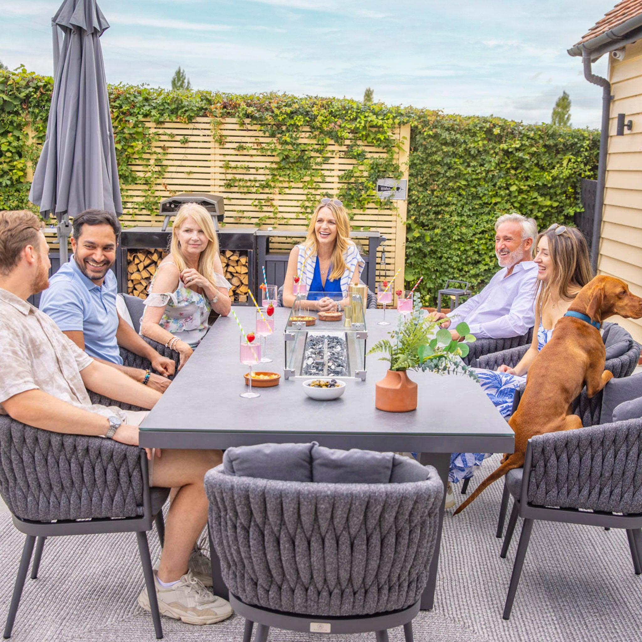 A group of six people and a dog sit around a Palma 8 Seat Rope Rectangular Ceramic Firepit Dining Set in Grey from Harbour Lifestyle, enjoying drinks and engaging in conversation. The Firepit Dining Set warms the evening as they all relax on UV-resistant chairs.