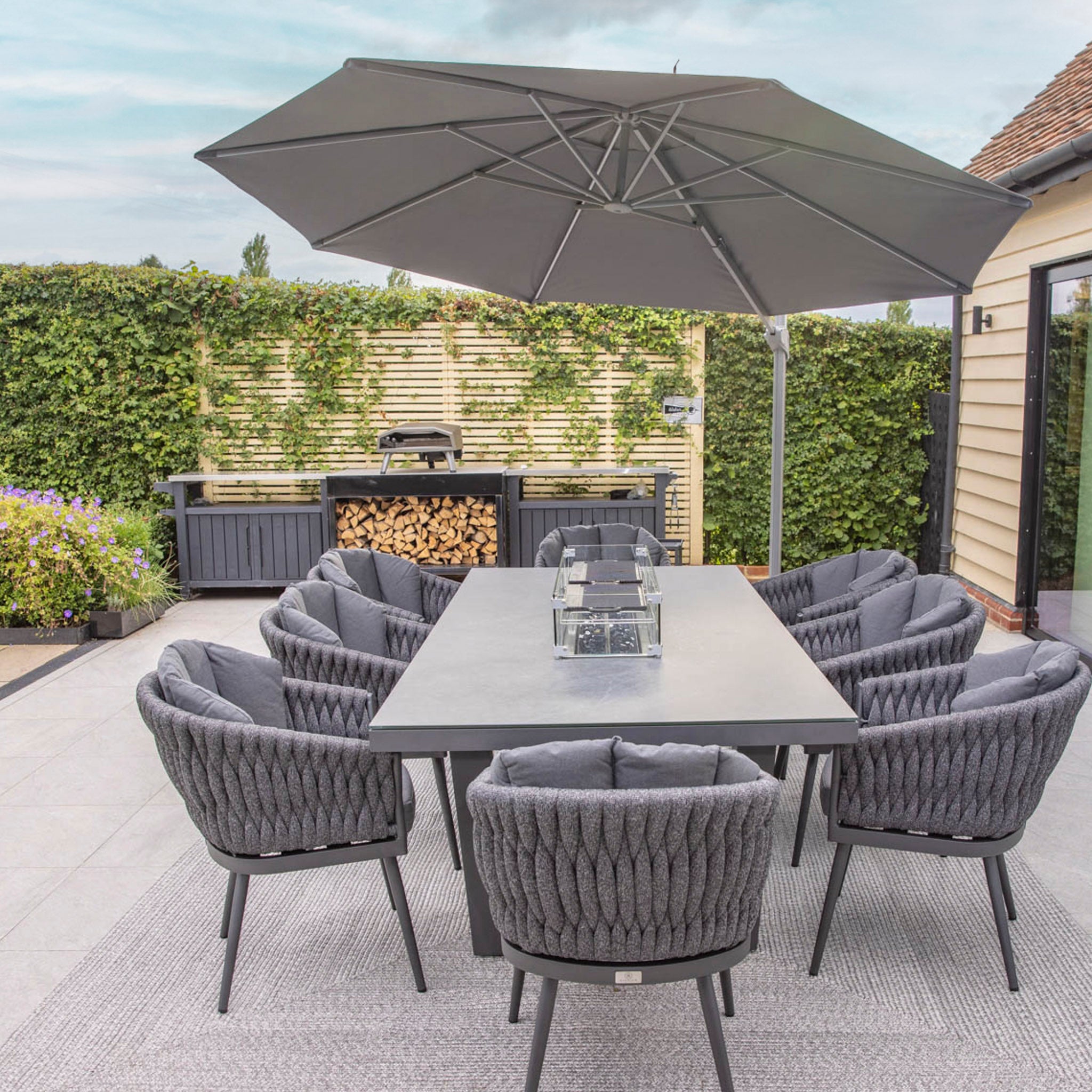 Modern outdoor dining area featuring a sleek Palma 8 Seat Rope Rectangular Ceramic Firepit Dining Set in Grey, with UV-resistant chairs with cushions, and a large umbrella, set on a patio near greenery.
