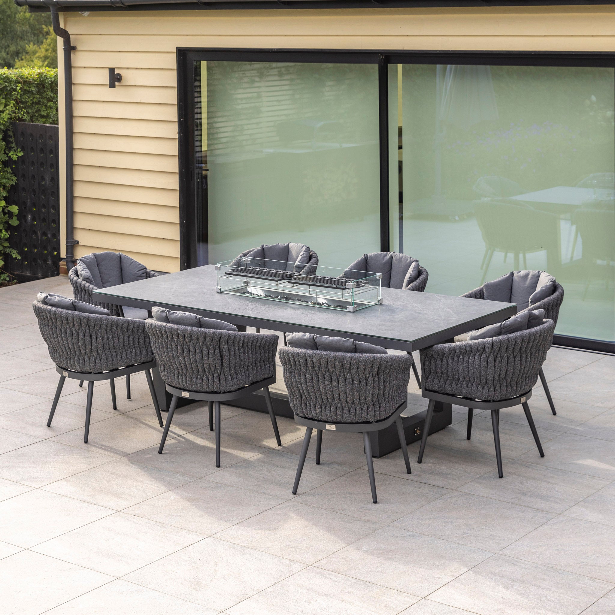 Outdoor patio featuring the Palma 8 Seat Rope Rectangular Ceramic Firepit Dining Set in Grey, with a modern dining table and eight UV-resistant cushioned chairs arranged around it on a stone-tiled surface.