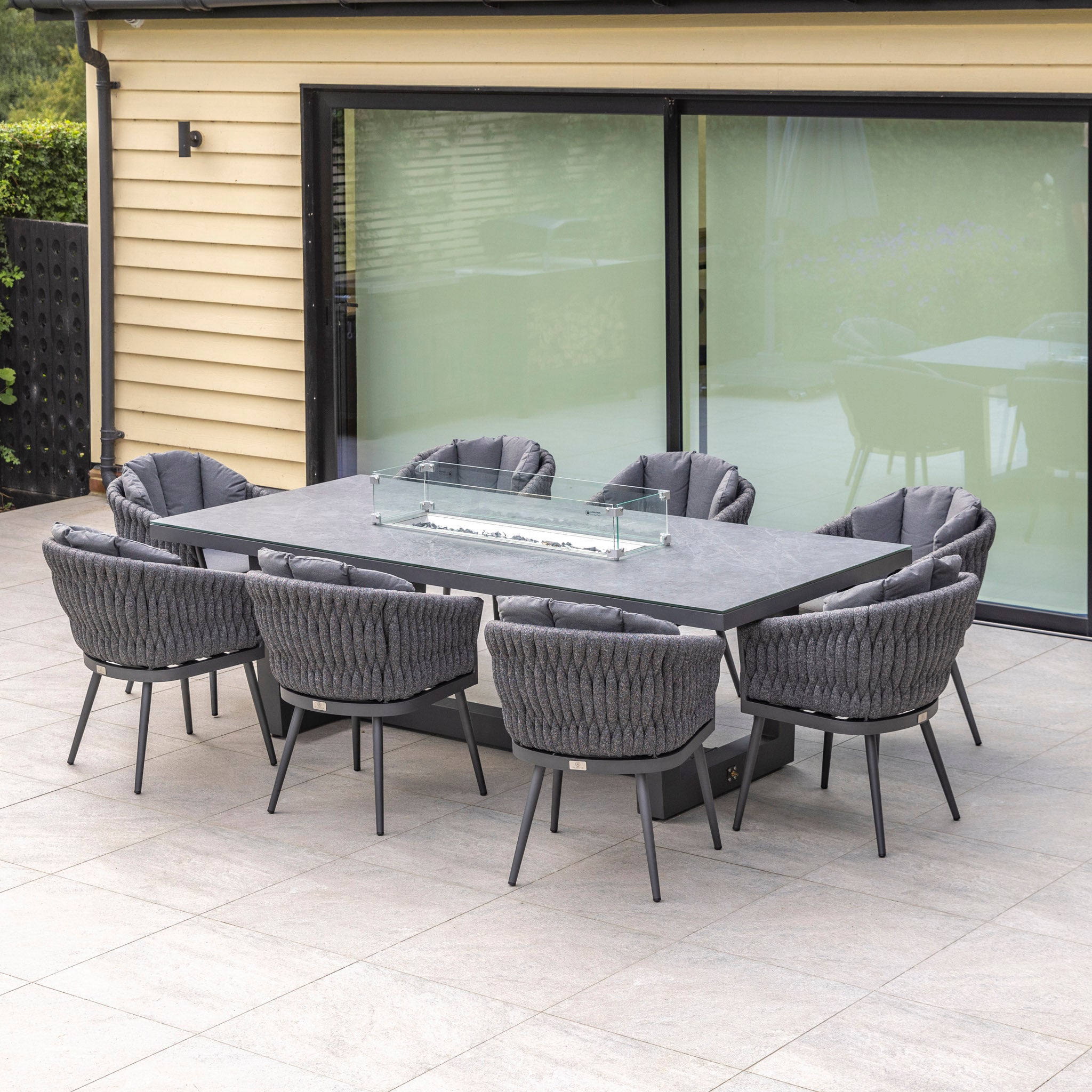 A contemporary dining experience with the Palma 8 Seat Rope Rectangular Ceramic Firepit Dining Set in Grey by Harbour Lifestyle, complete with eight UV-resistant cushioned gray chairs, perfectly situated on a patio near a sliding door.