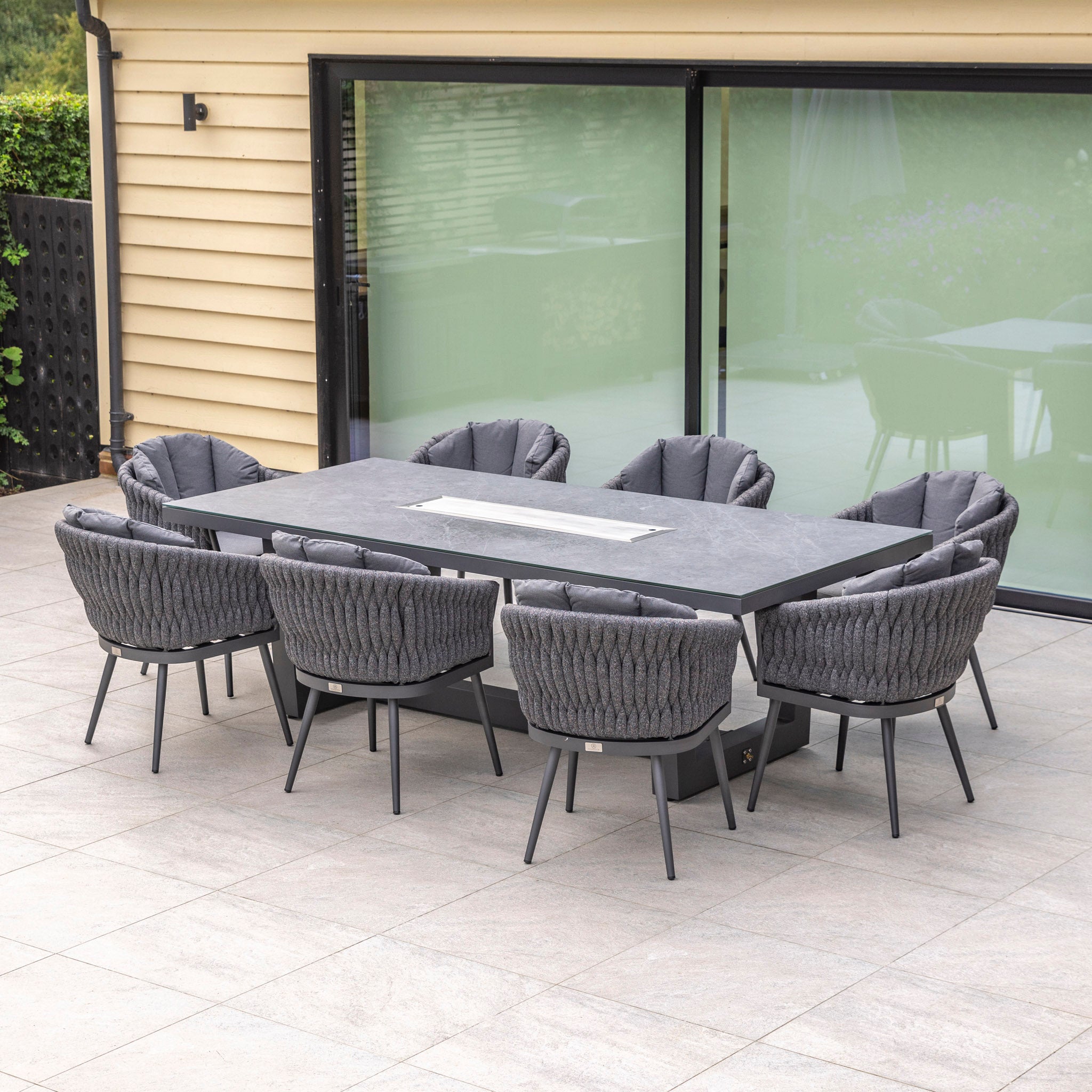 Outdoor dining area featuring a Palma 8 Seat Rope Rectangular Ceramic Firepit Dining Set in Grey, with a large rectangular table and eight UV-resistant cushioned chairs on a tiled patio in front of glass doors.