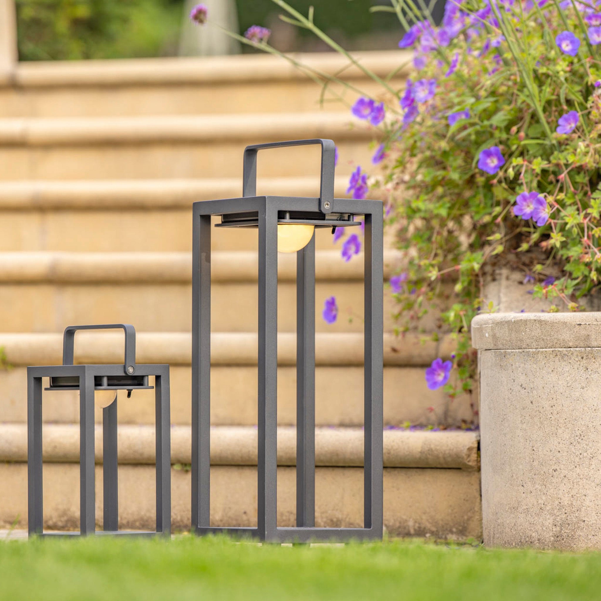 Two Luxor Large Floor Outdoor and Indoor Solar Lanterns in Charcoal sit on the grass near stone steps and a planter with blooming purple flowers. These weatherproof lights feature an automatic switch, making them perfect for any outdoor setting.