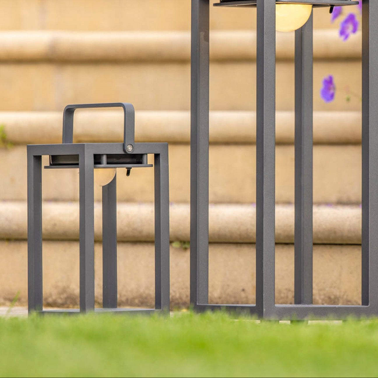 Two Luxor Small Table Outdoor and Indoor Solar Lanterns in charcoal provide mood lighting on the grass, with stone steps and purple flowers in the background.