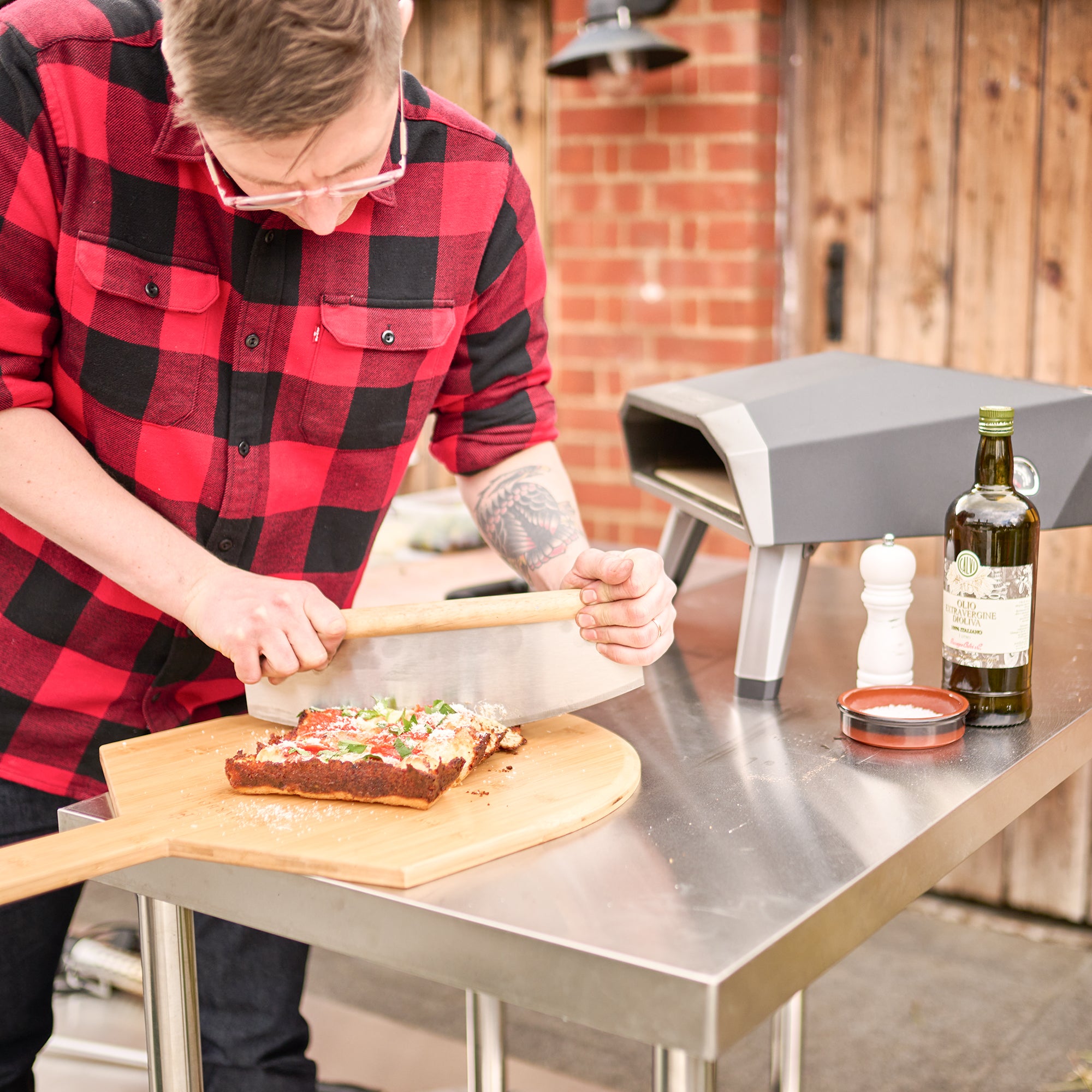 A person in a red checkered shirt is slicing pizza on a 16" Bamboo Pizza Board outside near an oven with condiments arranged on a metal table.