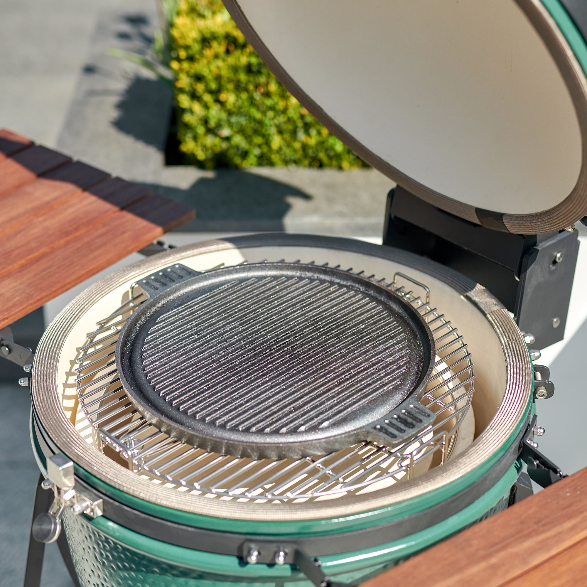 Open green ceramic grill with a Kamado Double Ear Cast Iron Plate griddle insert, wooden side tables, and a backdrop of plants.