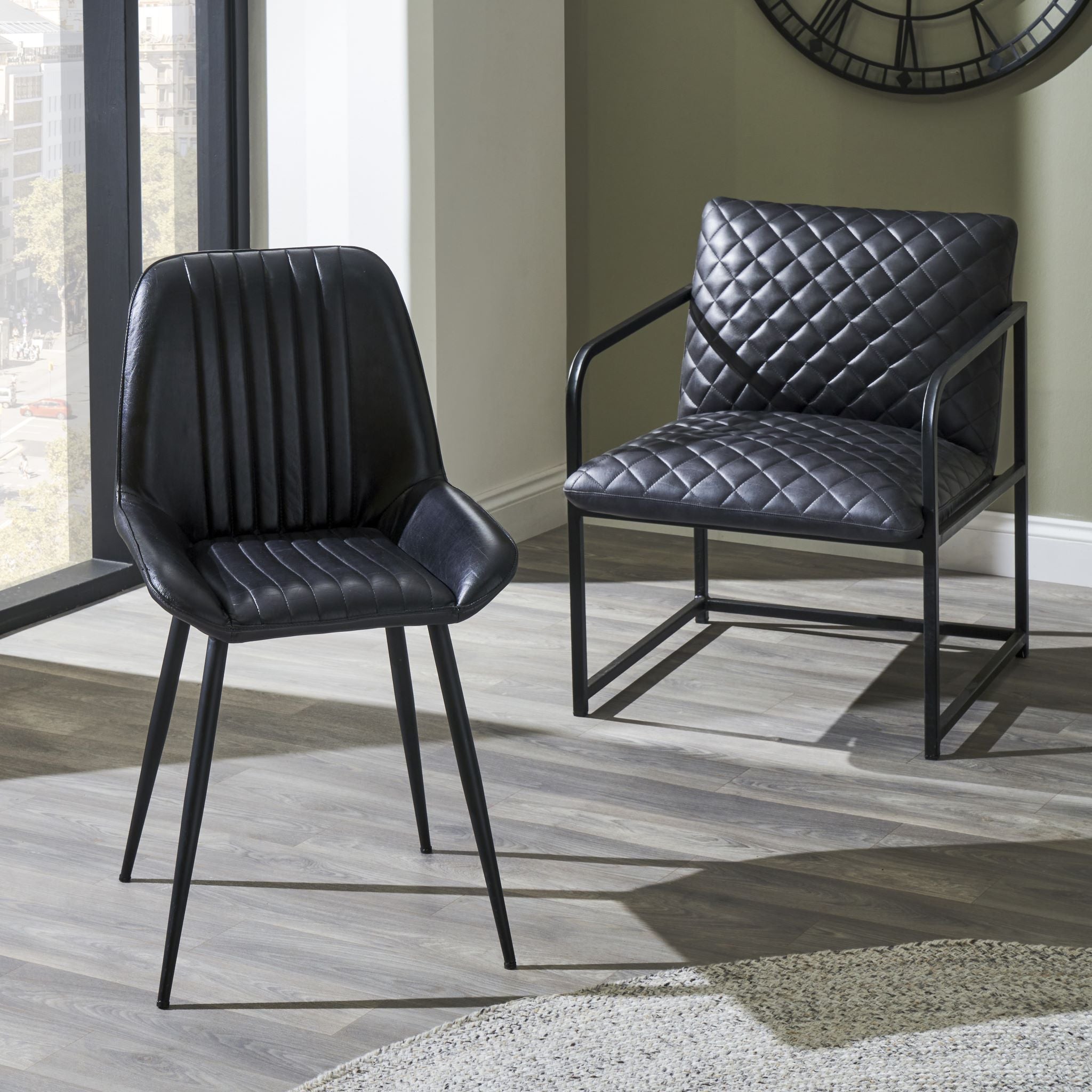 Two Pacific Lifestyle Angelo Leather and Iron Retro Dining Chairs in Steel Grey rest elegantly on a light wood floor, with a large window and wall clock in the background.