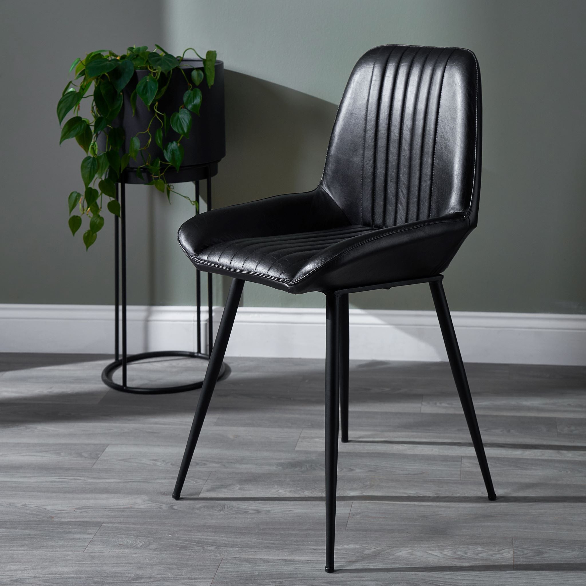 The Angelo Leather and Iron Retro Dining Chair in Steel Grey by Pacific Lifestyle elegantly rests on a wooden floor, enhanced by vertical stitching, with a potted plant on a stand nearby.