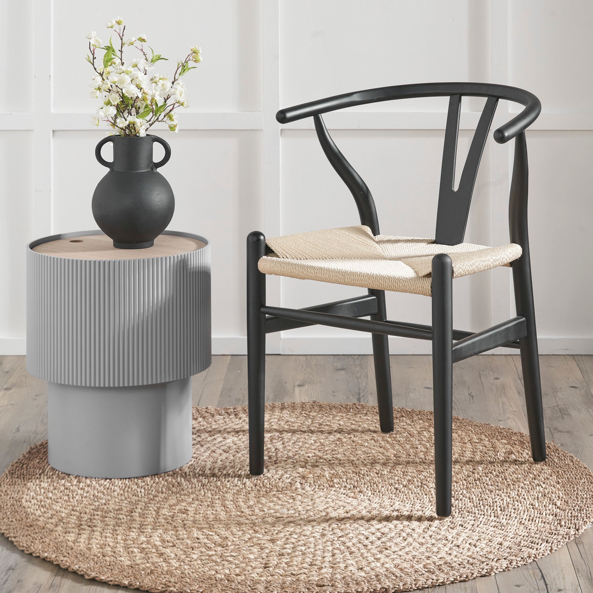 A Pacific Lifestyle Quinn Black Beech Wood and Natural Rope Dining Chair elegantly complements a small gray table. The set, adorned with a black vase and flowers, sits on a round natural rope rug, adding texture to the minimalist room.