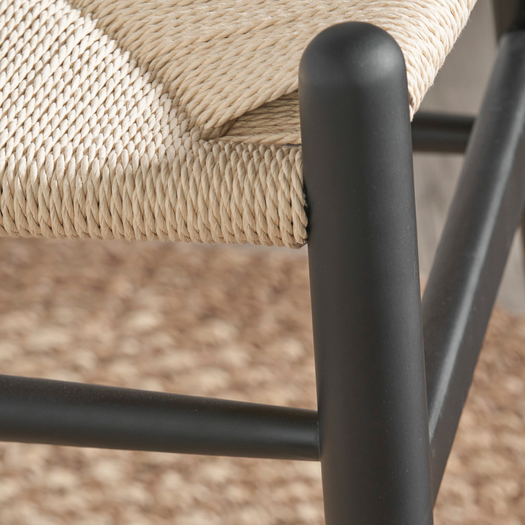 A close-up view of the Pacific Lifestyle Quinn Dining Chair showcases its woven beige seat made from natural rope, supported by a black cylindrical beech wood frame against a textured brown surface.