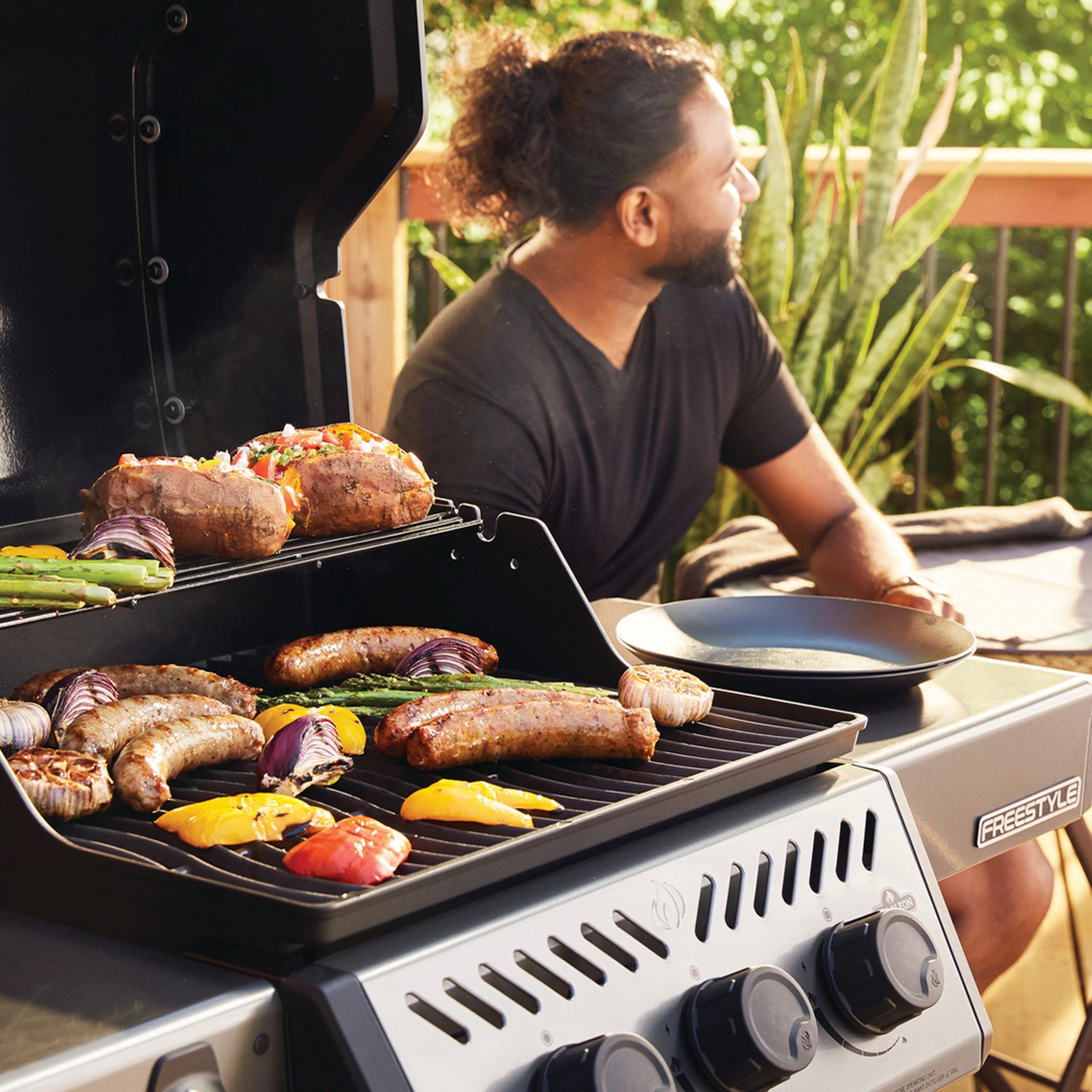 A man sits next to a Napoleon Freestyle 425 Gas Barbecue featuring a JETFIRE ignition system, grilling sausages, onions, and colorful bell peppers on cast iron porcelainized enamel cooking grids in a sunny, outdoor setting.