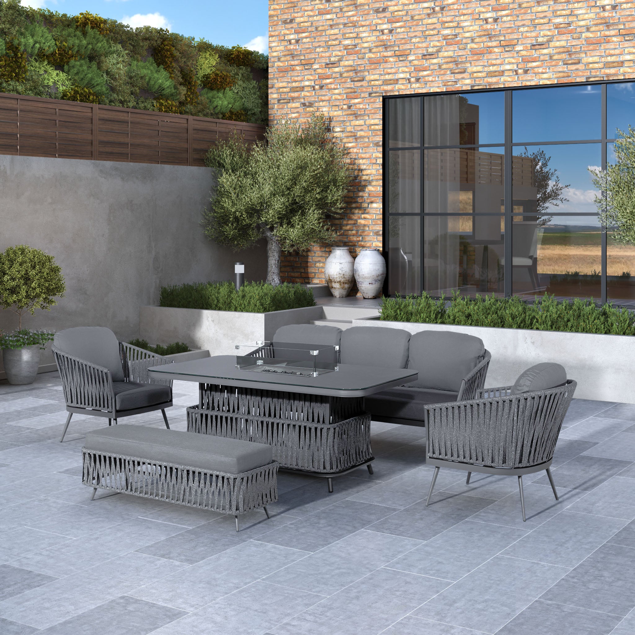 The Monterrey 3 Seat Rope Sofa Set with Rising Firepit Table in Grey from Harbour Lifestyle showcases modern gray woven furniture, a glass-top table with cushioned seats, and is perfect for placement in front of a brick building. This set also features a built-in rising firepit, adding both warmth and ambiance.