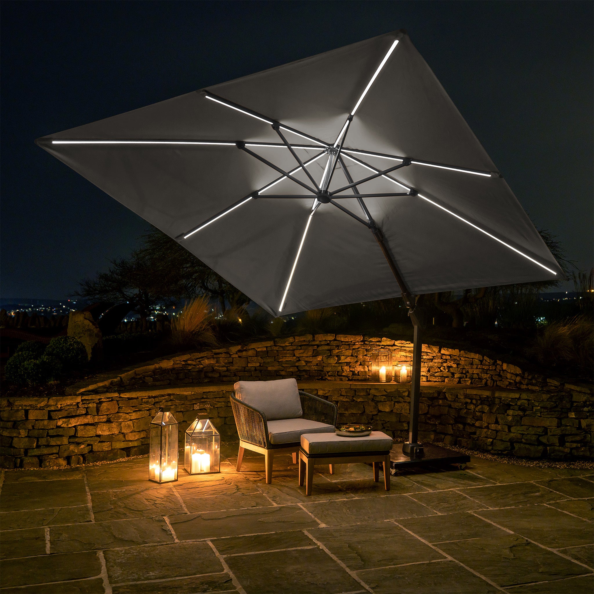 At night, a patio is illuminated by the Pacific Lifestyle Glow Challenger T2 3m Square Cantilever Parasol in Grey, two lanterns, a chair, and a table. Powder-coated aluminium complements the stone wall and floor for a cozy atmosphere.