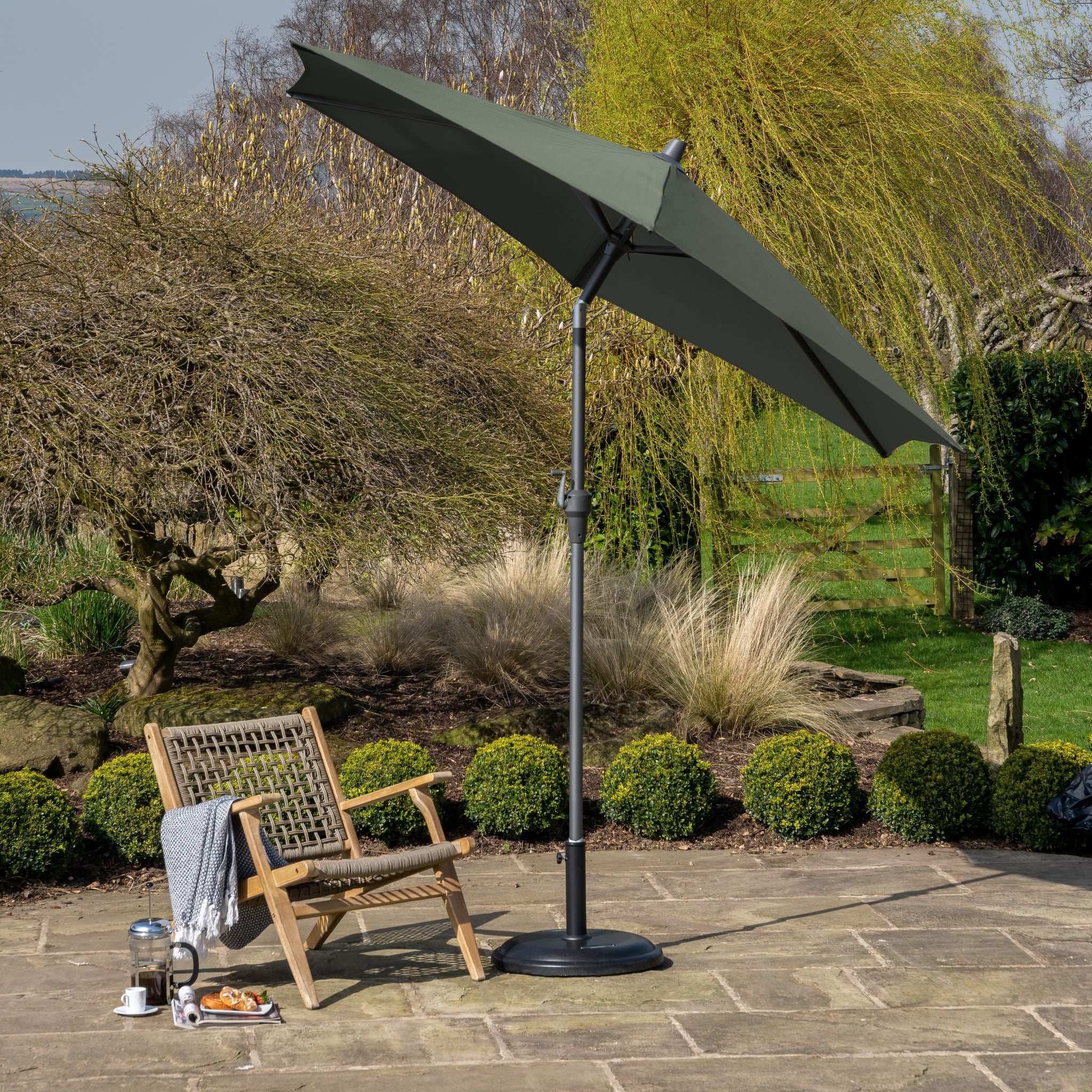 A serene patio features a Pacific Lifestyle - Riva 2.5m Round Green Parasol alongside a wooden chair draped with a blanket; the coffee set rests on stone amidst lush greenery.