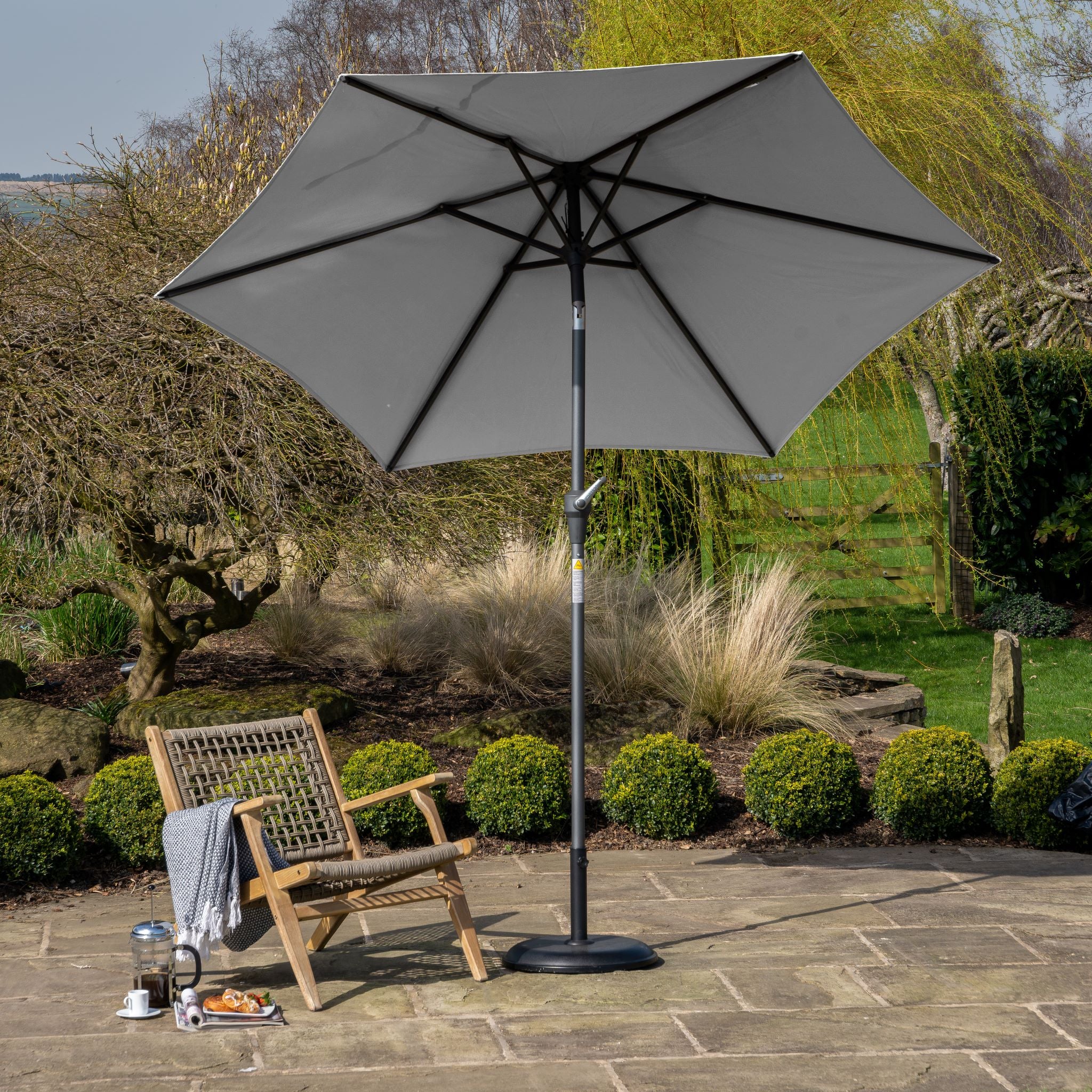 The patio showcases the Riva 3m Round Grey Parasol by Pacific Lifestyle with UV protection, alongside a wooden chair, cozy blanket, and mug. A powder-coated aluminum frame adds durability, while lush plants offer a serene backdrop.