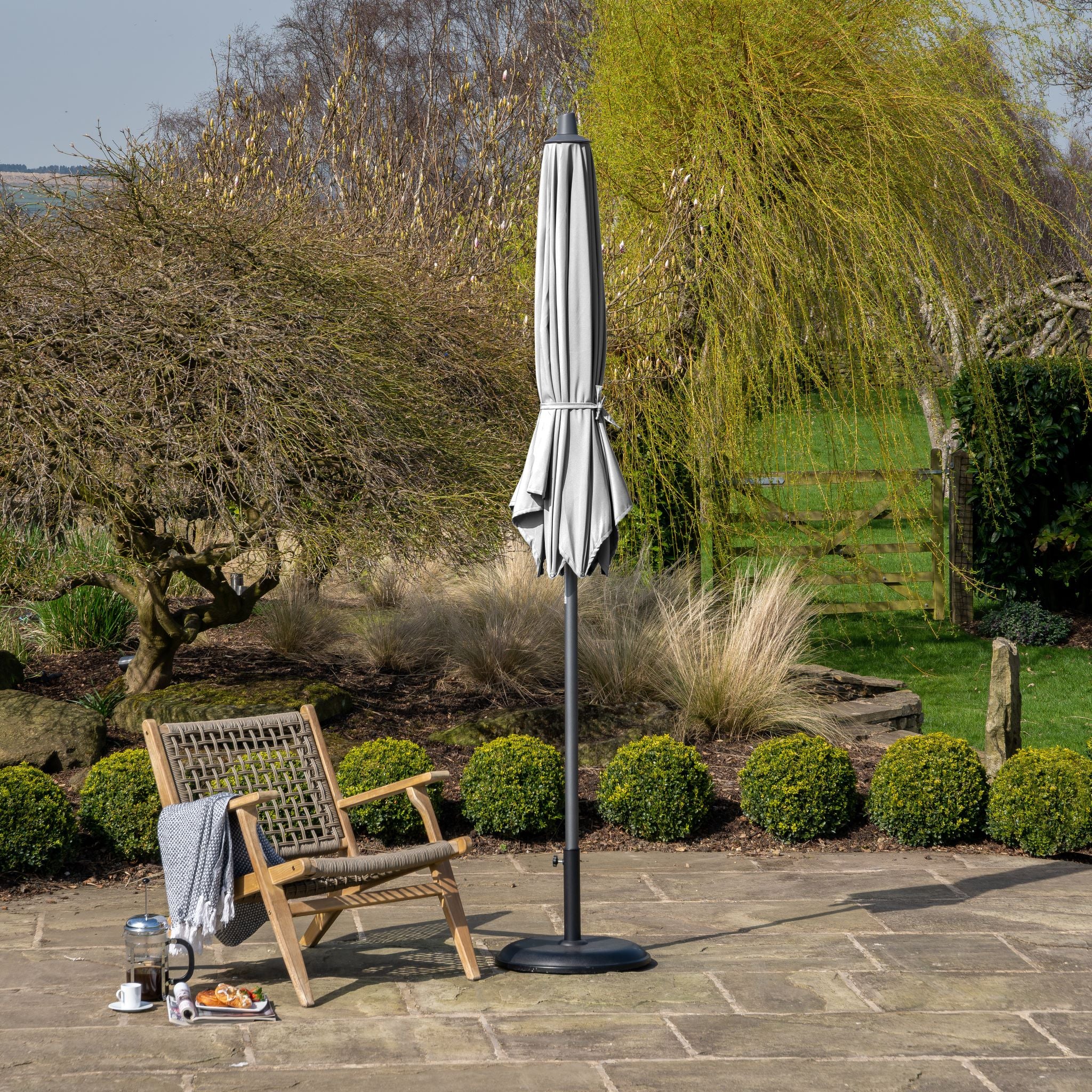 A patio with the Pacific Lifestyle Riva 3m Round Grey Parasol, featuring a powder-coated aluminum frame, stands over a wooden chair and tea set on stone, surrounded by lush greenery.