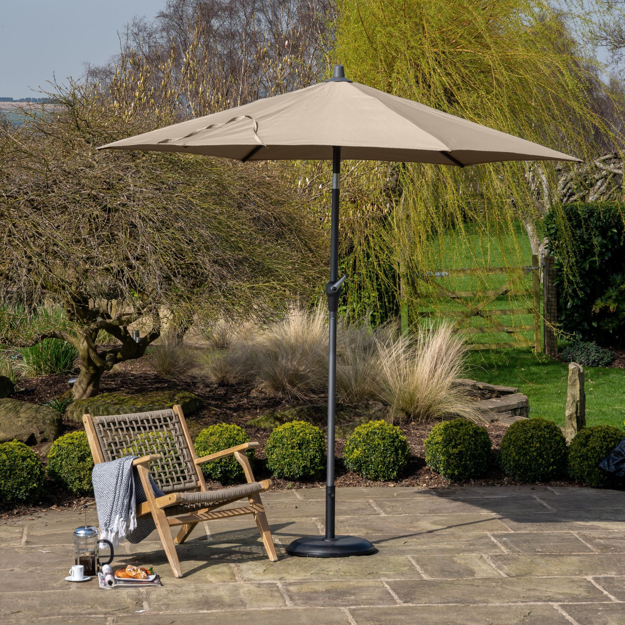 An outdoor patio with a wooden chair, table set with coffee and pastries, and the Pacific Lifestyle - Riva 3m Round Taupe Parasol featuring a powder-coated aluminium frame and durable fabric canopy, all surrounded by lush greenery.