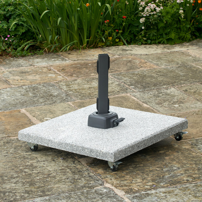 The Pacific Lifestyle Salerno Hammered Grey Granite 90KG Wheeled Parasol Base sits on a stone patio, surrounded by vibrant garden greenery and flowers.