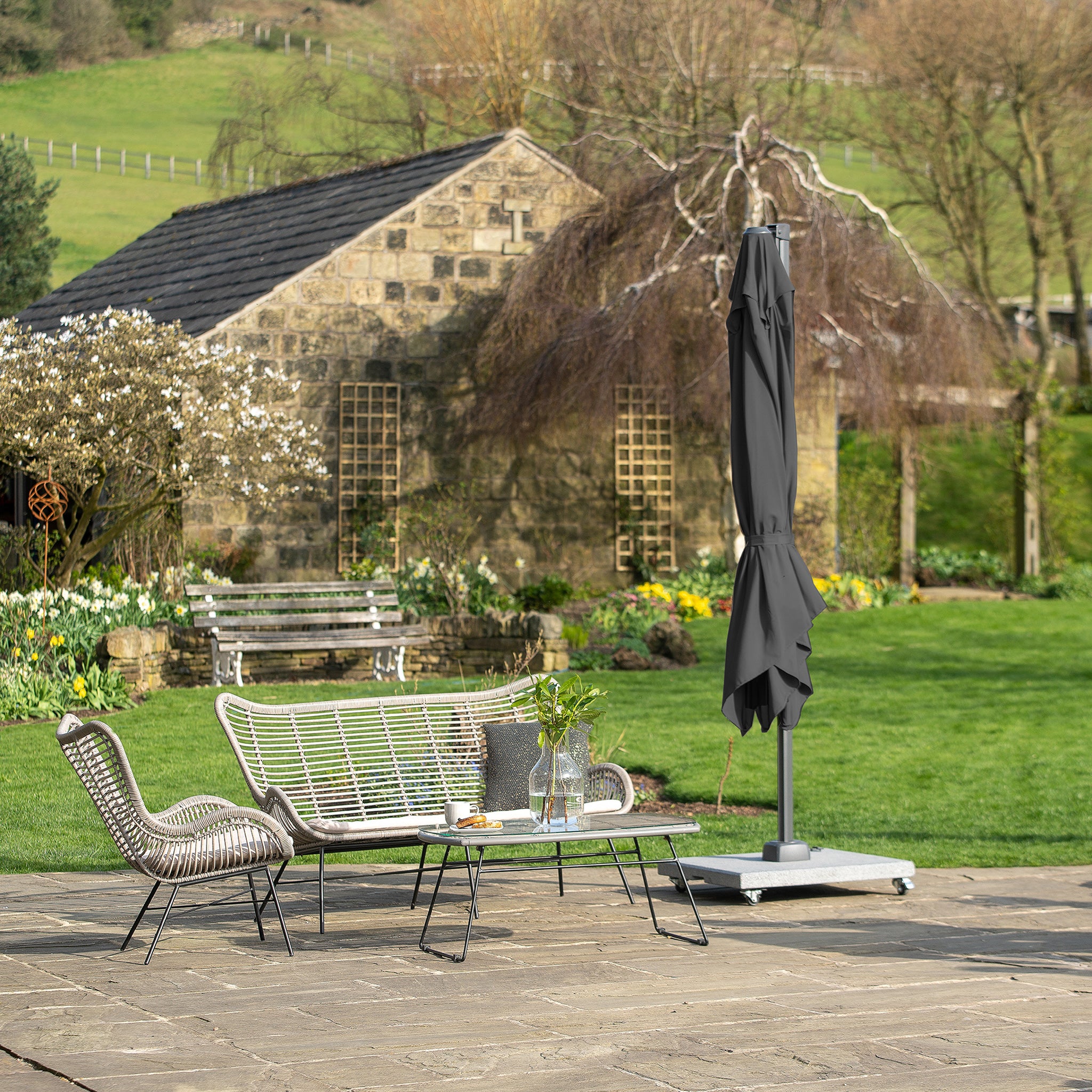 A charming patio with wicker furniture and a closed Pacific Lifestyle Voyager T2 2.7m Square Parasol in Anthracite enhances the wooden building in this lush garden setting.