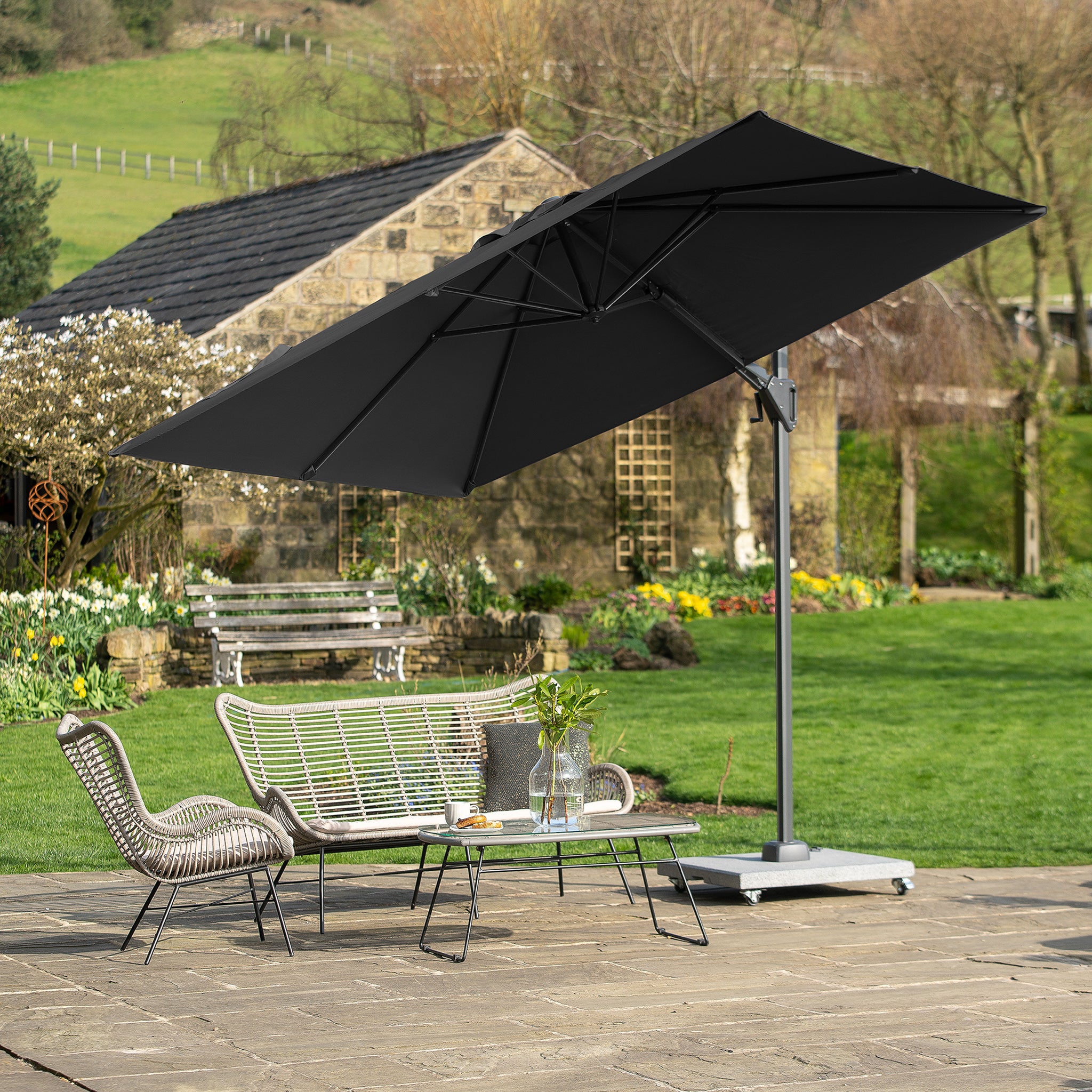 On a stone terrace, a patio furniture set is complemented by the Pacific Lifestyle Voyager T2 2.7m Square Parasol in Anthracite, all overlooking a lush green garden and rustic house.