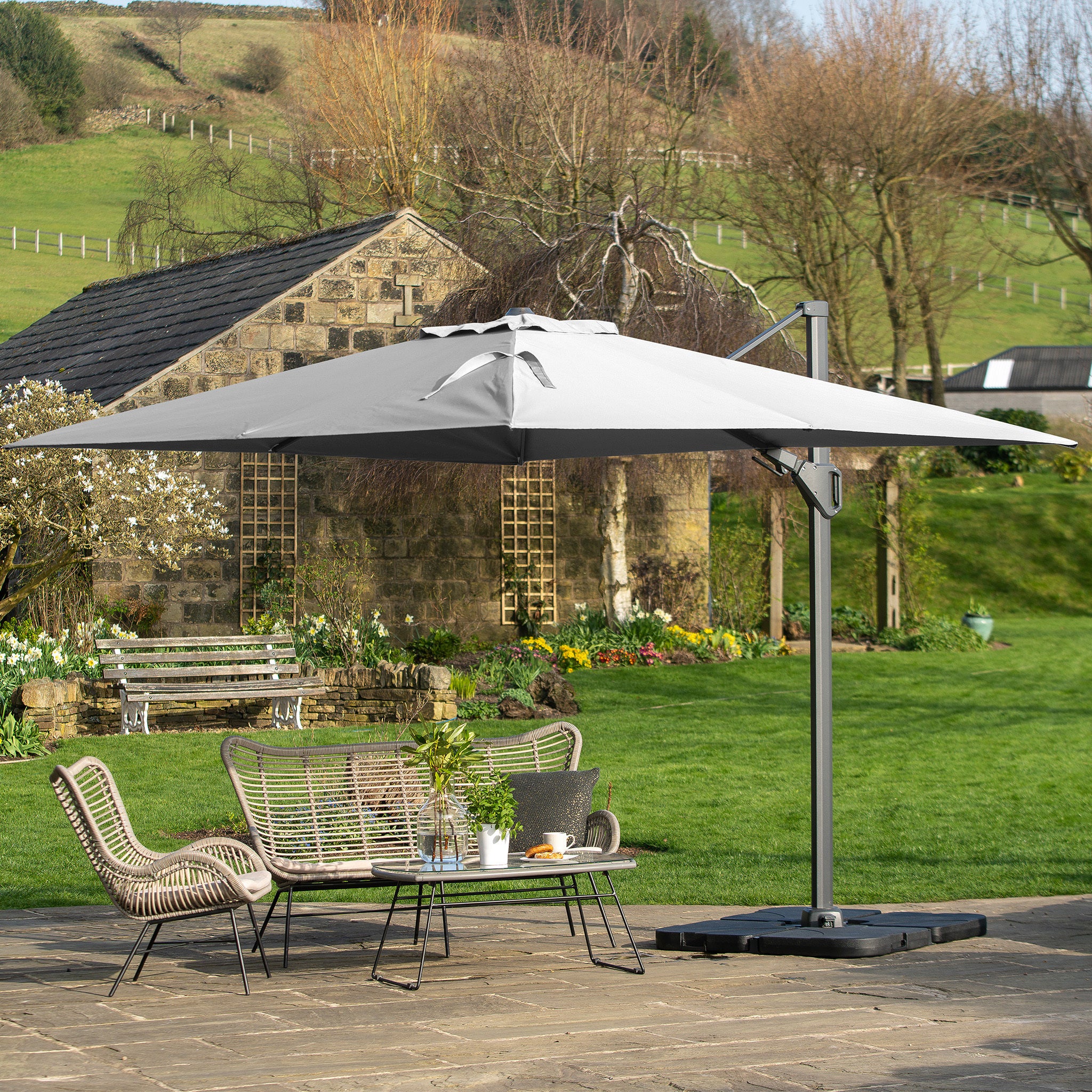 A charming patio showcases the Pacific Lifestyle Challenger T2 3.5m x 2.6m Rectangular Cantilever Parasol in Grey, complementing the wicker furniture and table set. A rustic cottage and lush garden create a serene backdrop, accented by durable powder-coated aluminum elements.