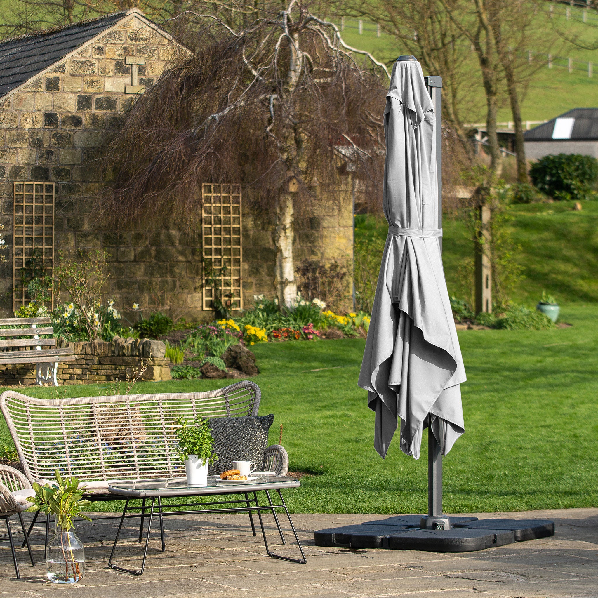 A garden patio features a closed Pacific Lifestyle Challenger T2 Rectangular Cantilever Parasol in Grey, wicker chairs, and a small table near a stone building.