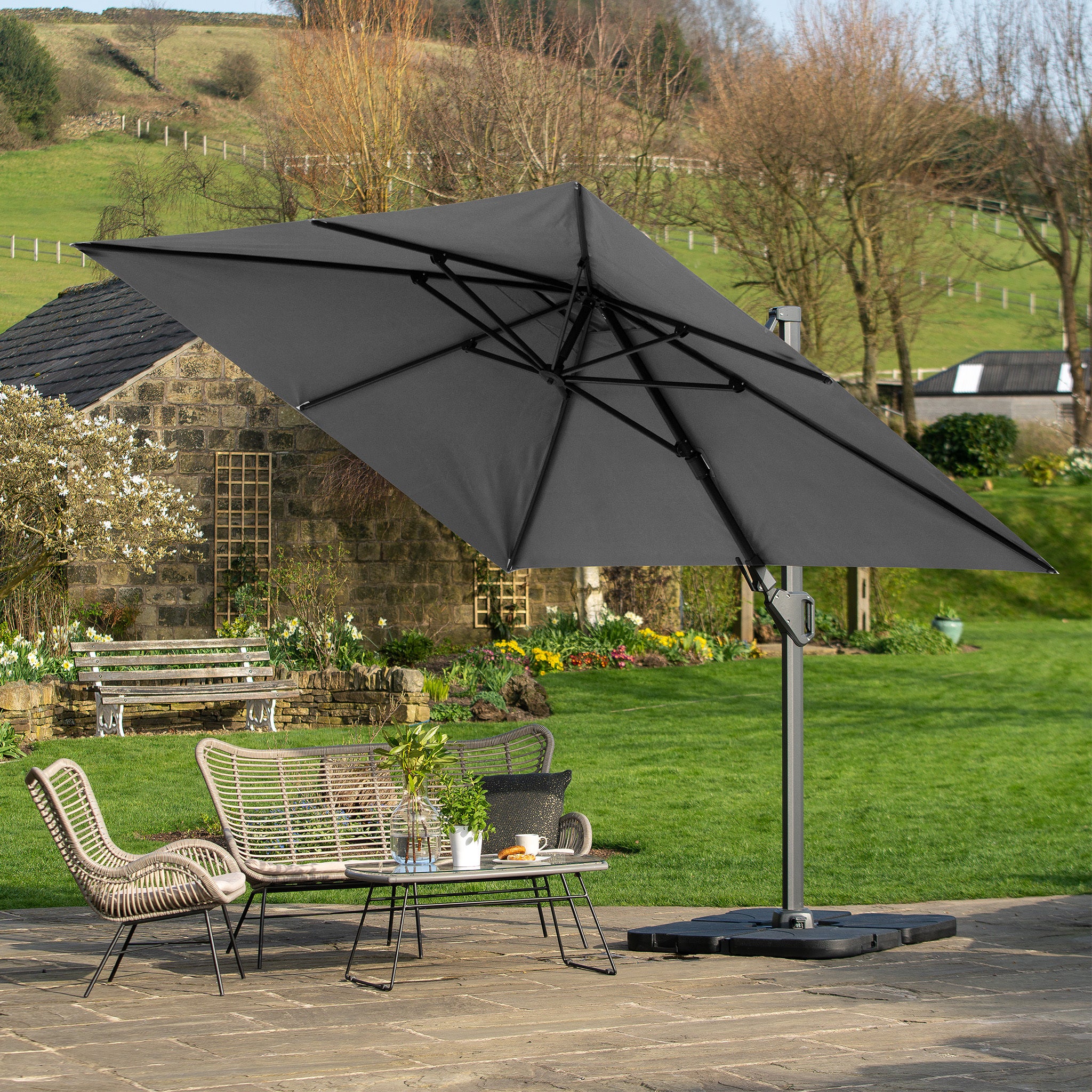 A Pacific Lifestyle - Challenger T2 3.5m x 2.6m Grey Rectangular Cantilever Parasol with a sleek double tilt system elegantly covers outdoor furniture on the stone patio. Its powder-coated aluminium frame enhances the serene garden and historic stone building, adding style and durability.