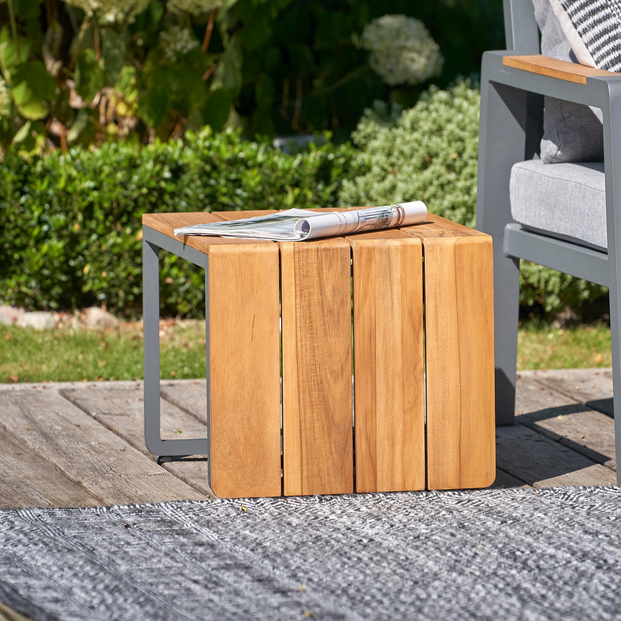 A wooden outdoor side table with teak detailing supports a rolled-up newspaper, complementing the Pacific Lifestyle Stockholm 3 Seat Sofa Set in marl grey and a gray cushioned chair on the patio.