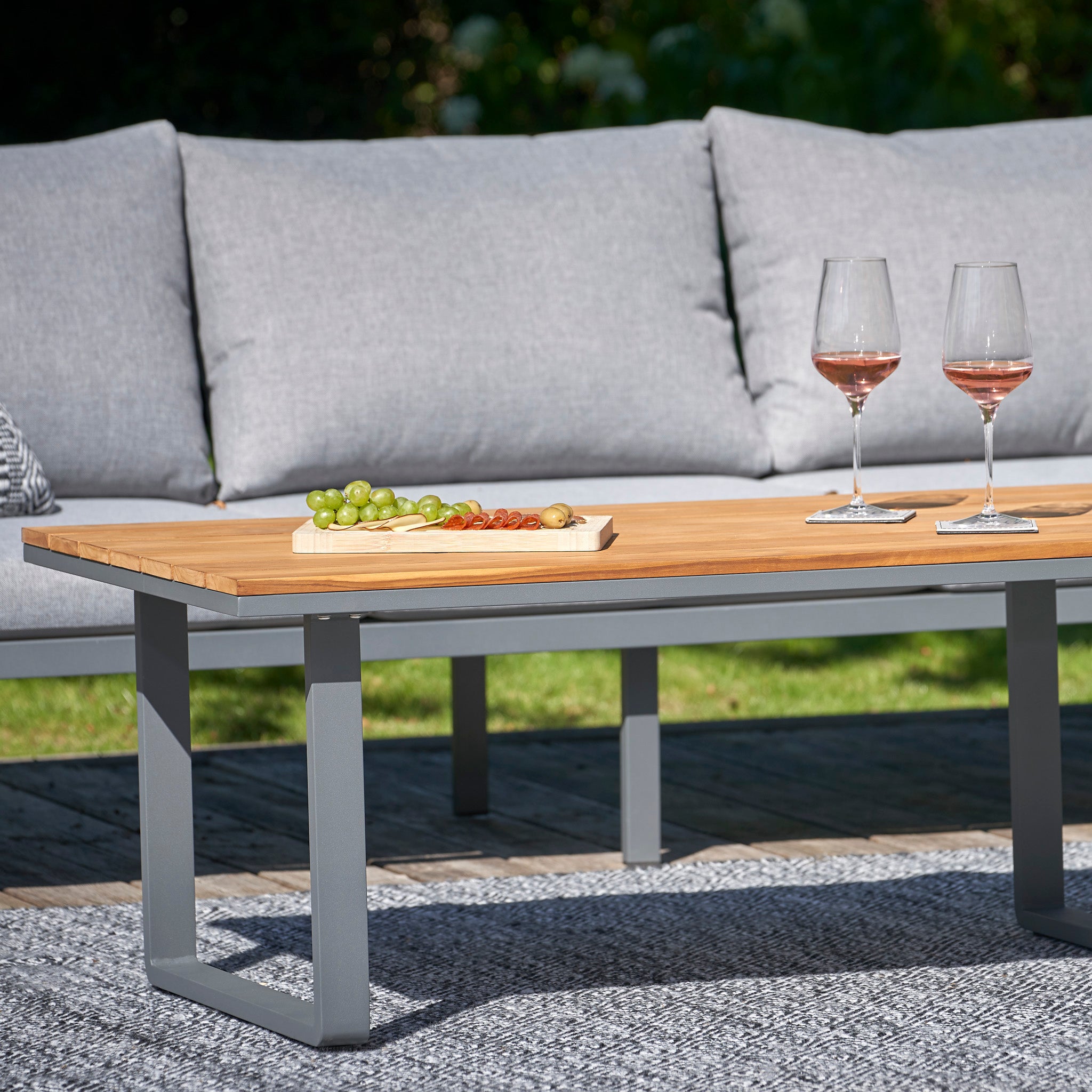 The outdoor seating area showcases the Pacific Lifestyle Stockholm 3 Seat Sofa Set in Marl Grey with teak accents, alongside a wooden table topped with wine glasses, grapes, and snacks.