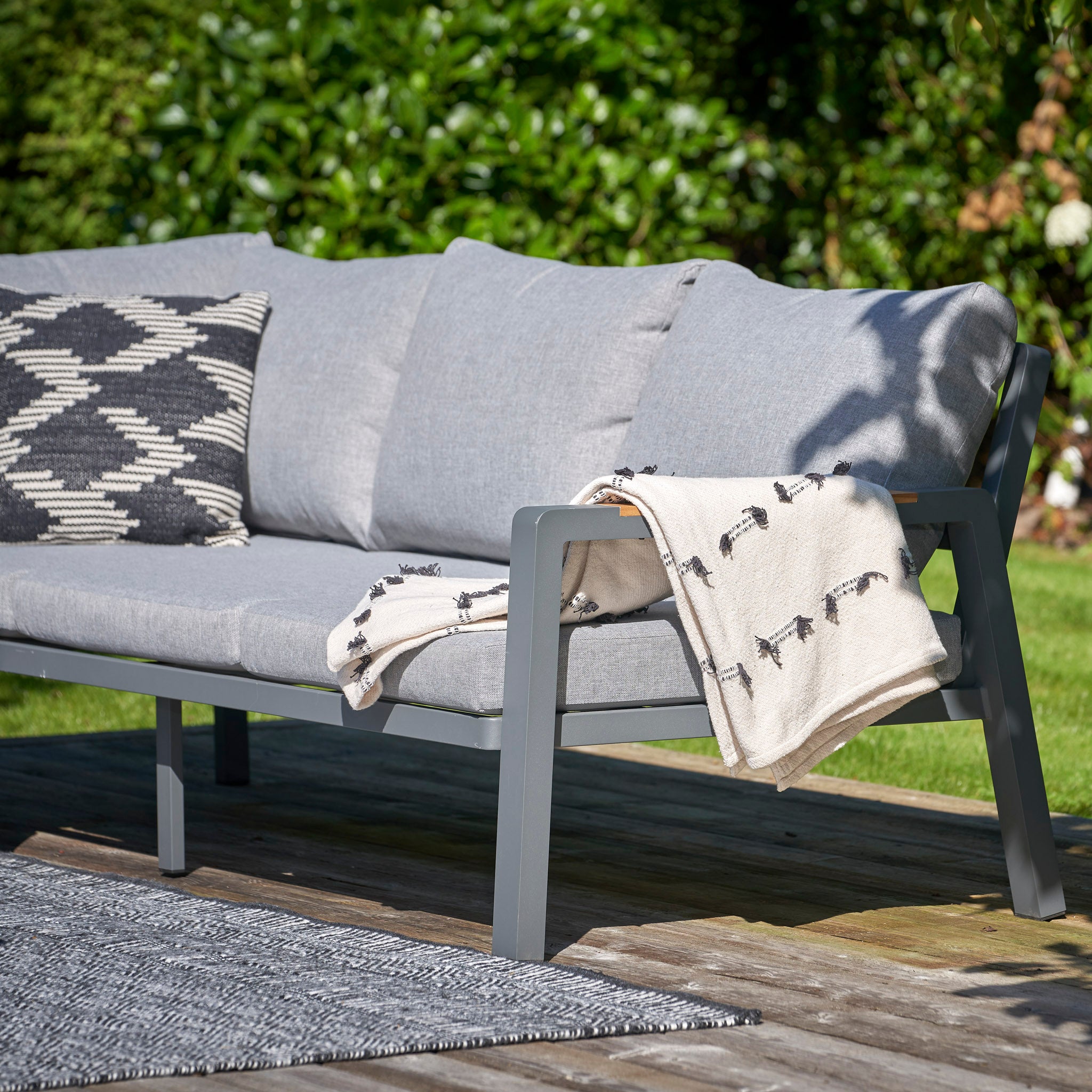 The Stockholm 3 Seat Sofa Set in Marl Grey by Pacific Lifestyle, adorned with pillows and a patterned blanket, is arranged on a wooden deck with teak detailing amidst lush greenery.