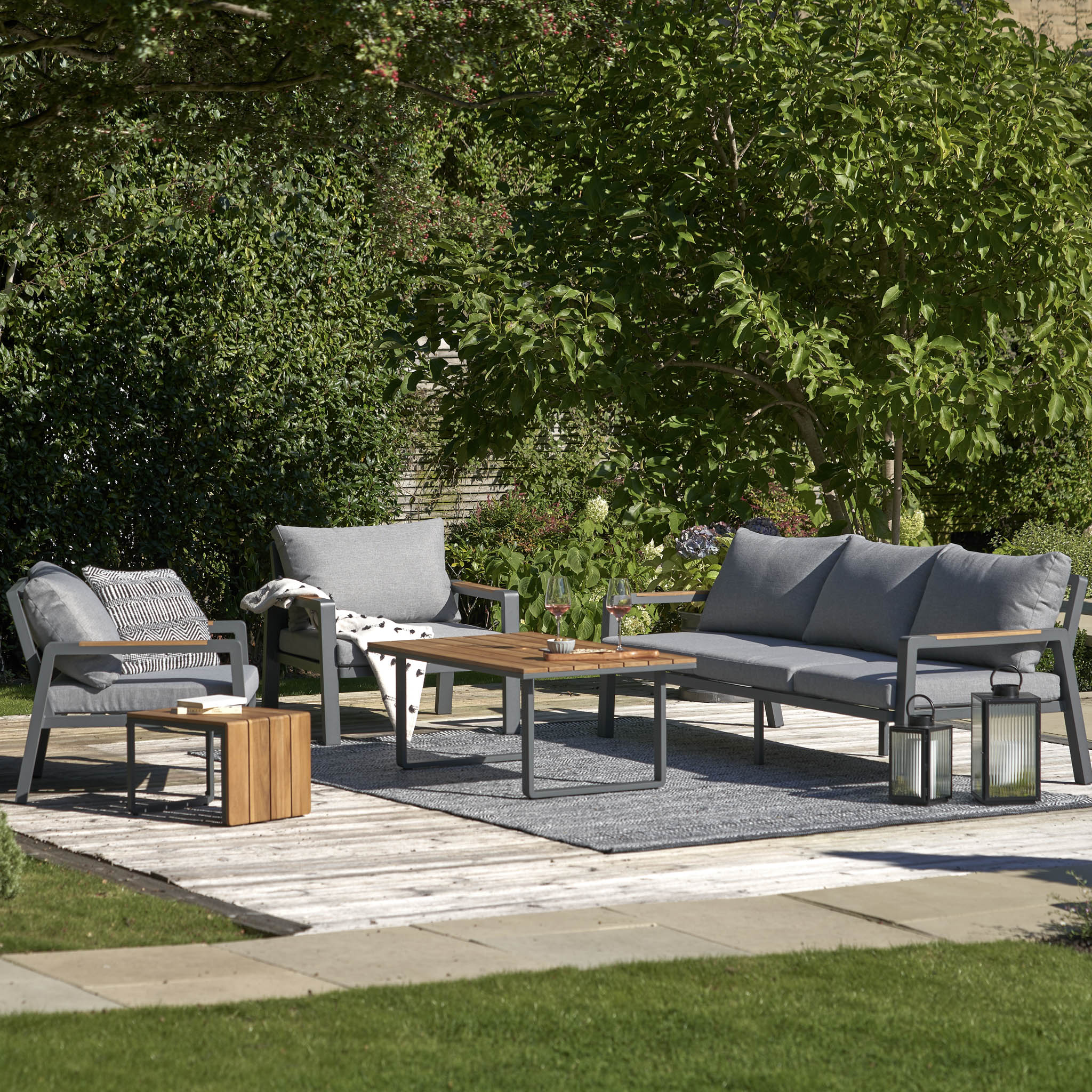 The Pacific Lifestyle - Stockholm 3 Seat Sofa Set in Marl Grey includes comfy gray couches and a wooden table with Teak Wood detailing. Paired with a cozy rug, elegant lanterns, and lush greenery, it creates an ideal outdoor relaxation spot.