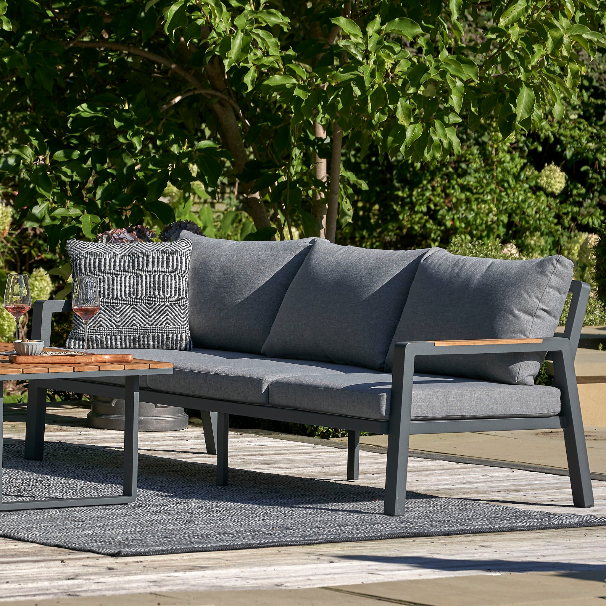The Stockholm 3 Seat Sofa Set in Marl Grey by Pacific Lifestyle showcases subtle teak wood details, perfect for patios with lush greenery and tree backdrops.