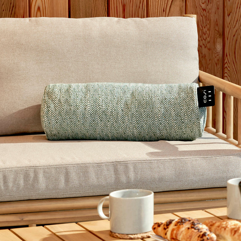 A cozy scene features the Cosi - Cosipillow Heated Bolster in Green on a beige outdoor sofa, with coffee cups and pastries invitingly arranged on a wooden table.