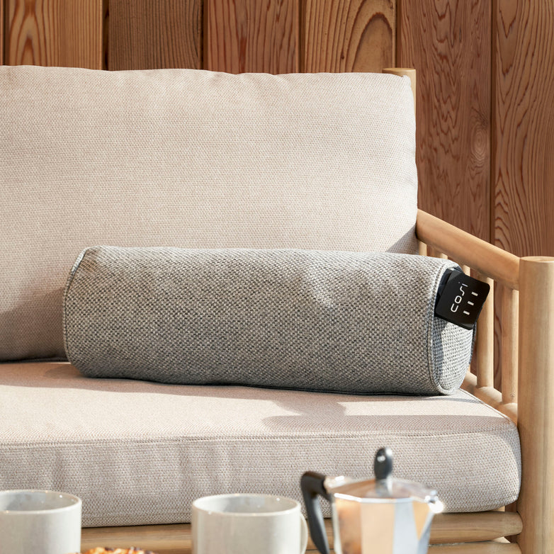 A Cosi Cosipillow Heated Bolster in grey adorns a beige sofa with a wooden backrest, while a coffee set on the small table nearby invites relaxation.