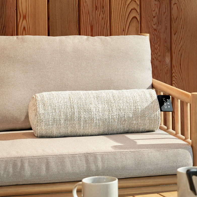 A beige sofa sits outdoors with the Cosi - Cosipillow Heated Bolster in Natural, joined by mugs on a table and an infrared heating element radiating nearby warmth.