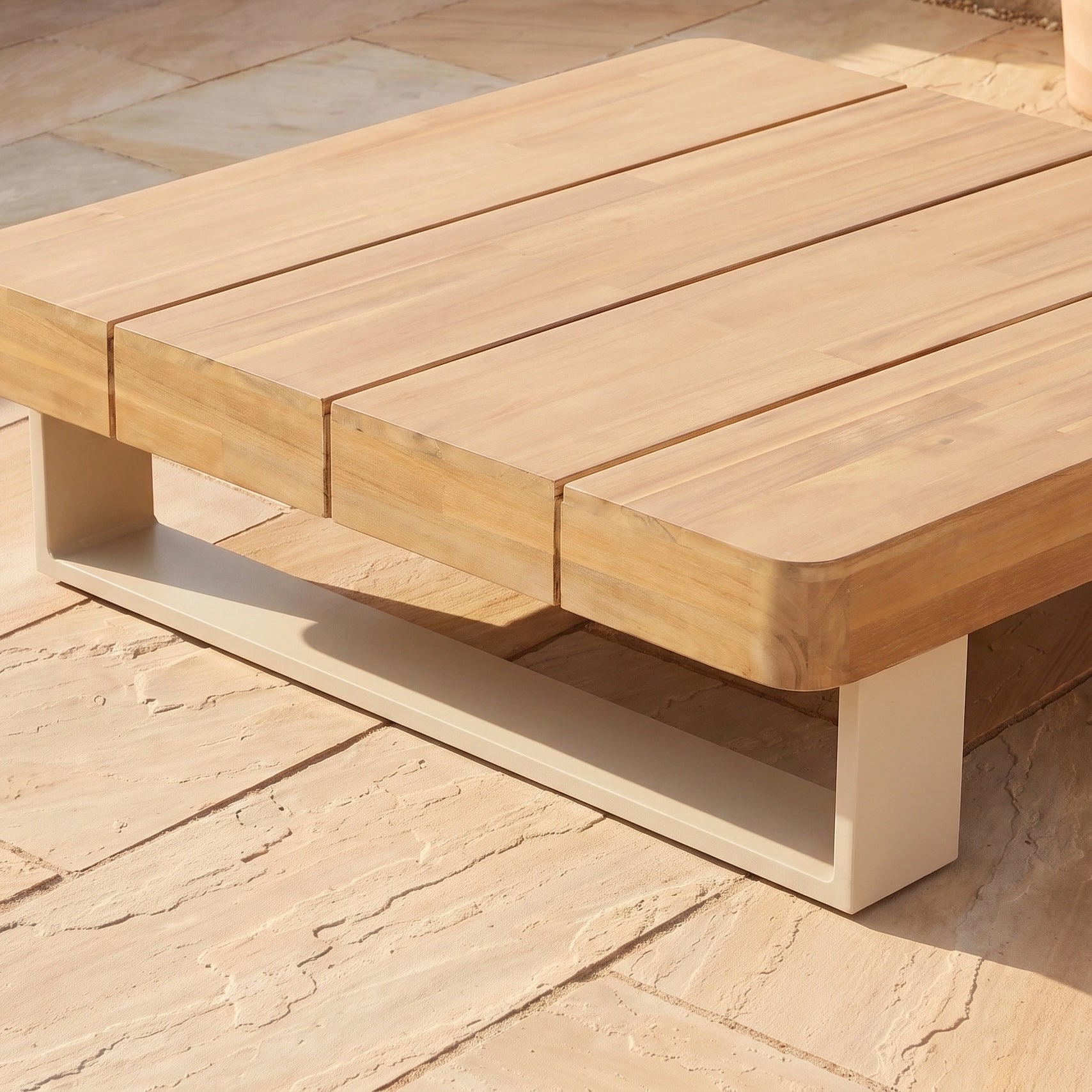 A low wooden coffee table, crafted from sustainable acacia wood with wide slats, sits gracefully on a beige stone patio. Sunlight casts soft shadows, enhancing the inviting atmosphere and complementing the surrounding outdoor furniture—perfectly paired with the Ava Corner Sofa Set in Latte.