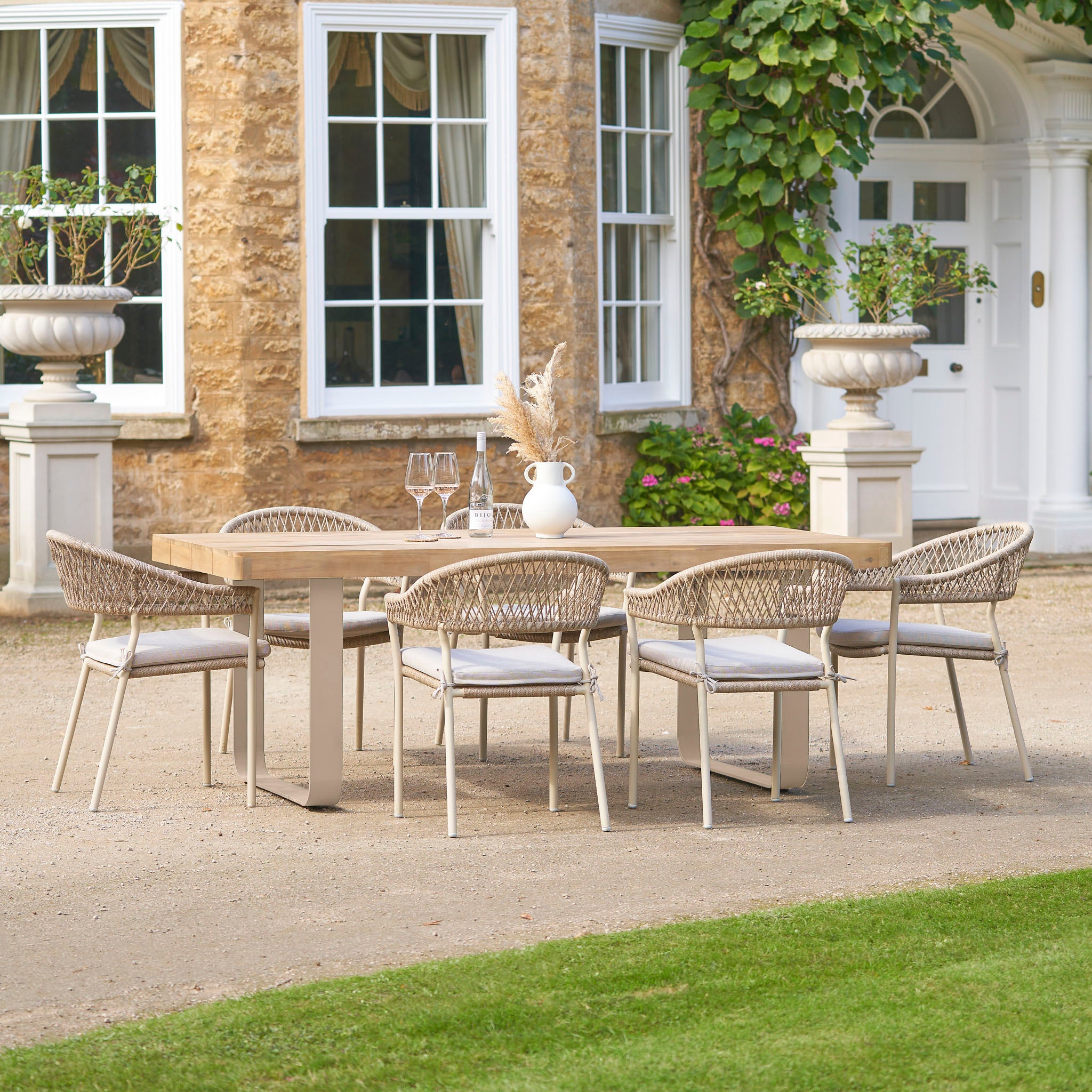 The Pacific Lifestyle - Osaka 6 Seat Dining Set in Latte, featuring six chairs and an Acacia wood table, is ideal for stylish patio gatherings in front of a stone house with large windows.