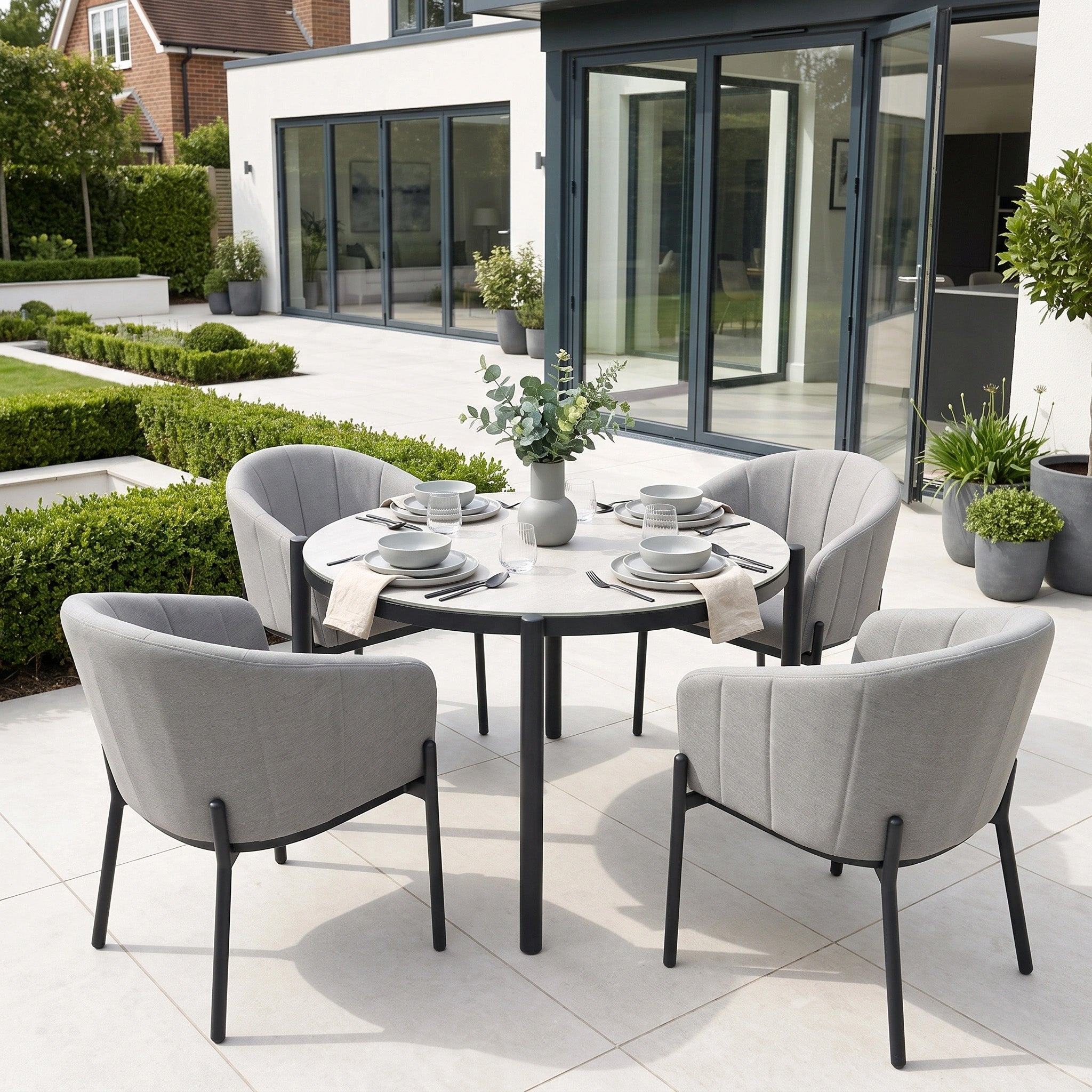 The Kai 4 Seat Round Dining Set in Light Grey is arranged on a modern patio beside a glass-walled house—perfect for outdoor dining with friends or family.