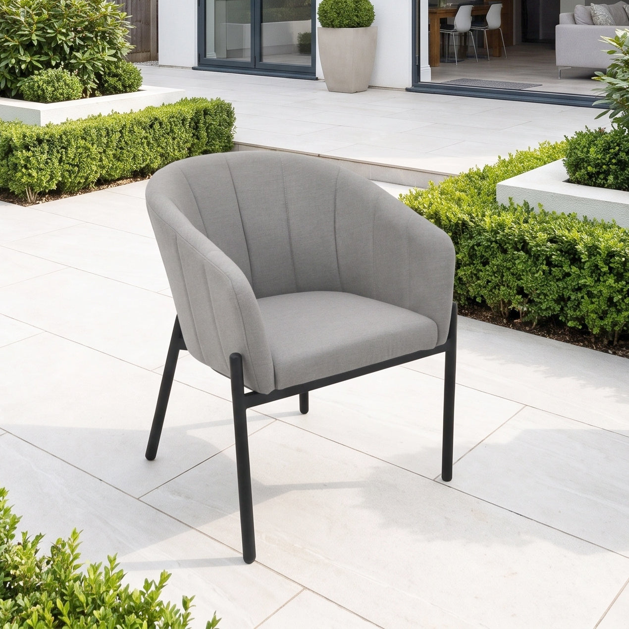 The Kai 4 Seat Round Dining Set in Light Grey sets the perfect tone for any patio—this gray cushioned chair with black legs blends beautifully among green shrubs and modern outdoor decor, making it an ideal centerpiece for stylish alfresco dining.