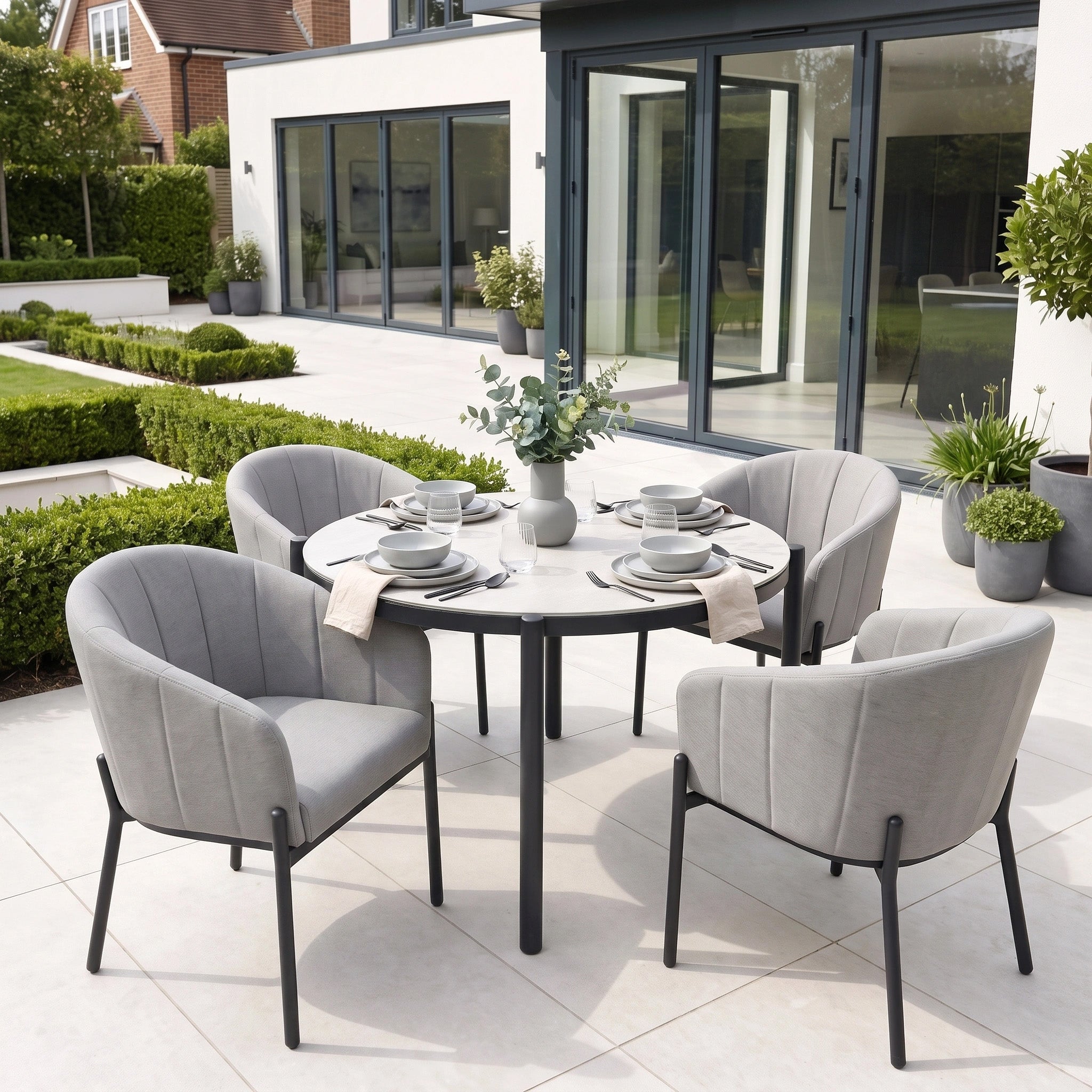 A modern patio featuring the Kai 4 Seat Round Dining Set in Light Grey with a stylish round table, four sleek light grey garden chairs, and elegant tableware, set outside a contemporary house—perfect for relaxed outdoor dining.