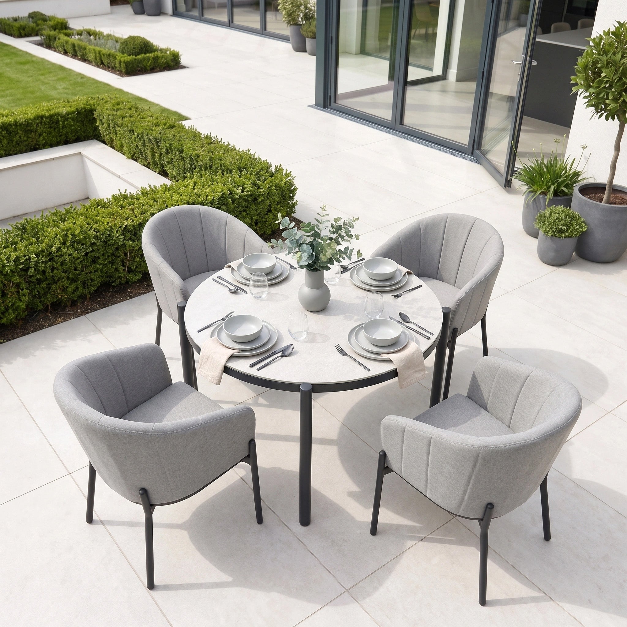 The Kai 4 Seat Round Dining Set in Light Grey, featuring a round patio table and four matching chairs, creates a chic and comfortable setup for alfresco dining on a contemporary tiled terrace accented with lush greenery. This stylish set effortlessly blends comfort with elegance, making it the ideal choice for outdoor gatherings.