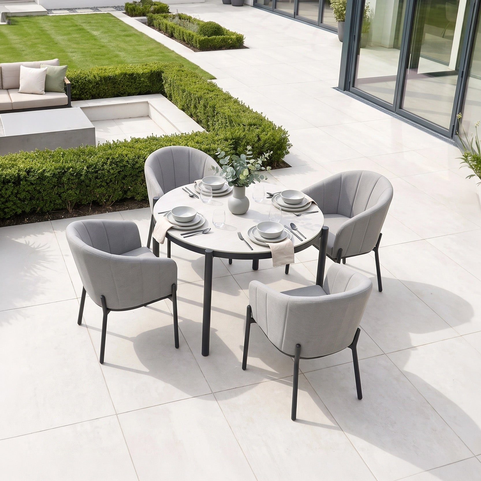 Modern outdoor patio featuring the Kai 4 Seat Round Dining Set in Light Grey, complete with four gray chairs and tableware arranged on white tile flooring. This elegant set of light grey garden furniture creates a stylish and inviting space for al fresco gatherings.