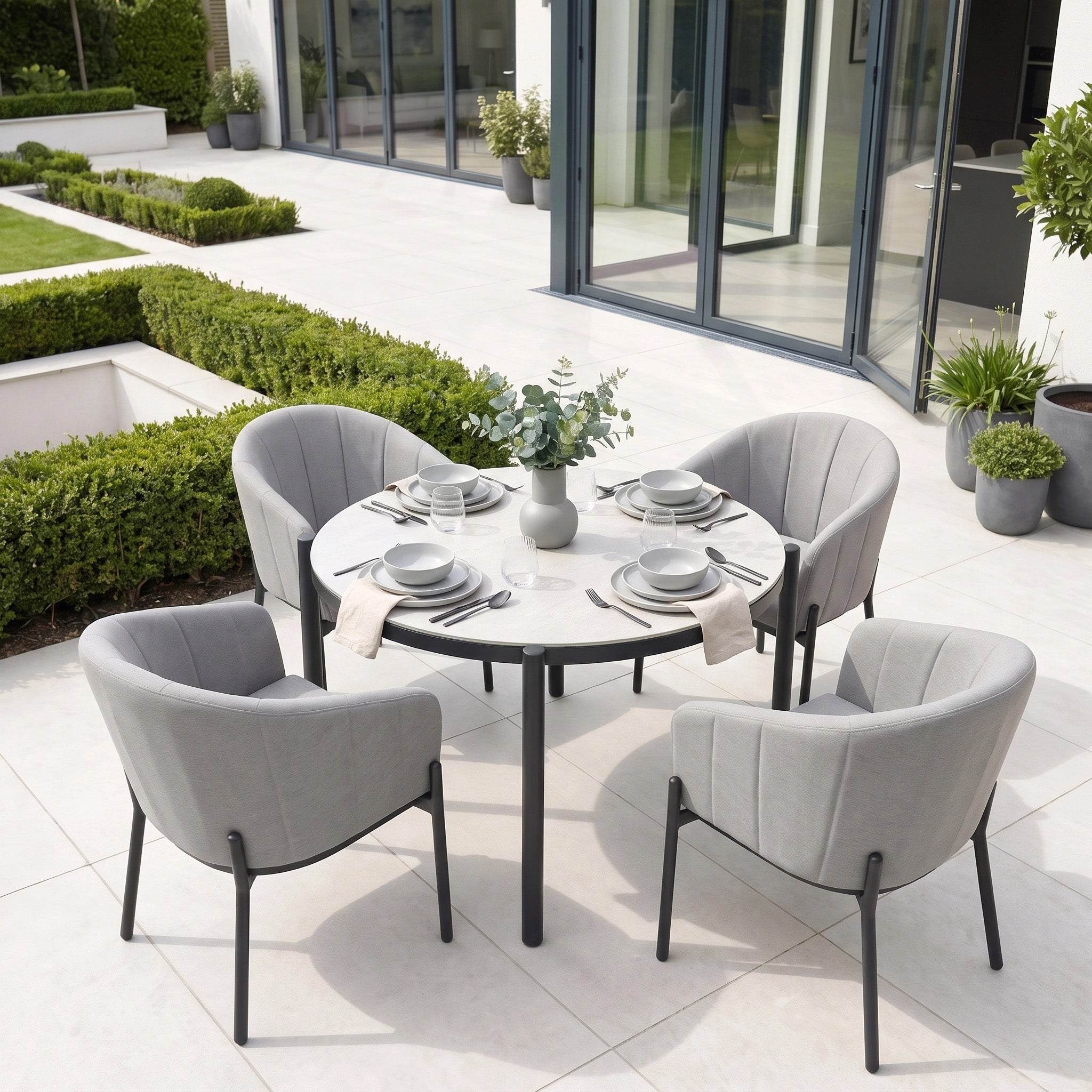 Modern outdoor patio featuring the Kai 4 Seat Round Dining Set in Light Grey, surrounded by gray chairs and lush green plants.
